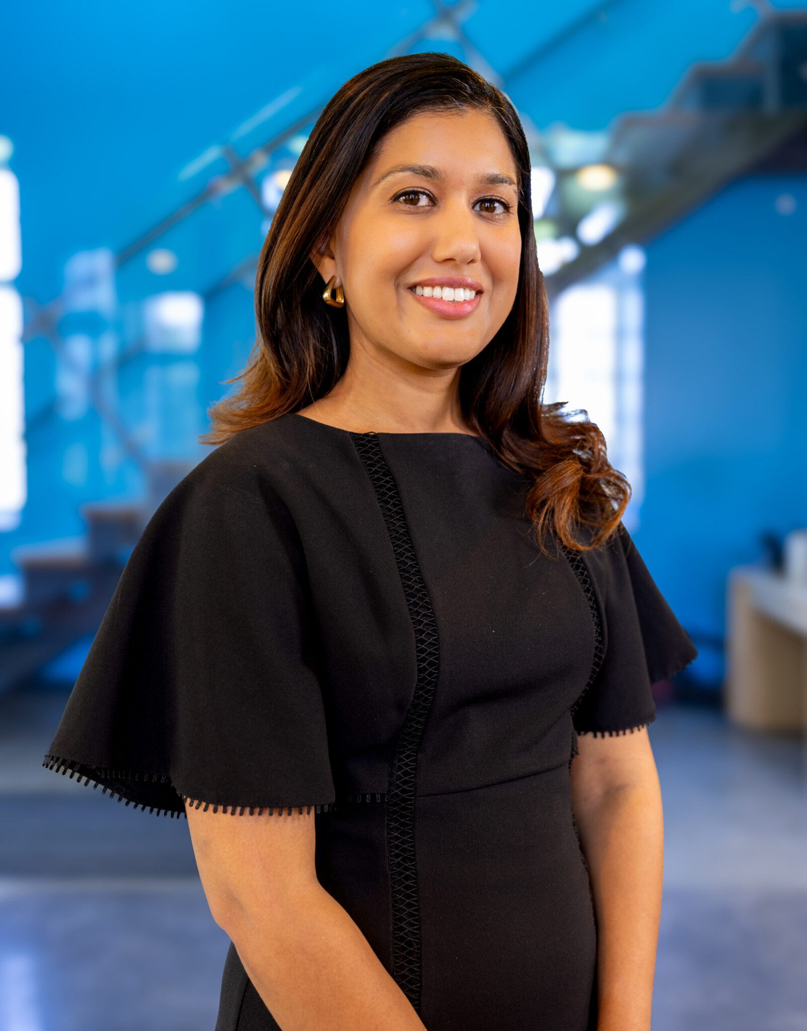 A woman with long brown hair, wearing a black dress, stands smiling in front of a modern indoor background with blue walls and a staircase.