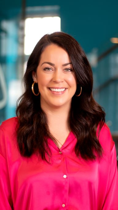 A woman with long dark hair, wearing gold hoop earrings and a bright pink button-up shirt, smiles while standing indoors against a blurred teal background.