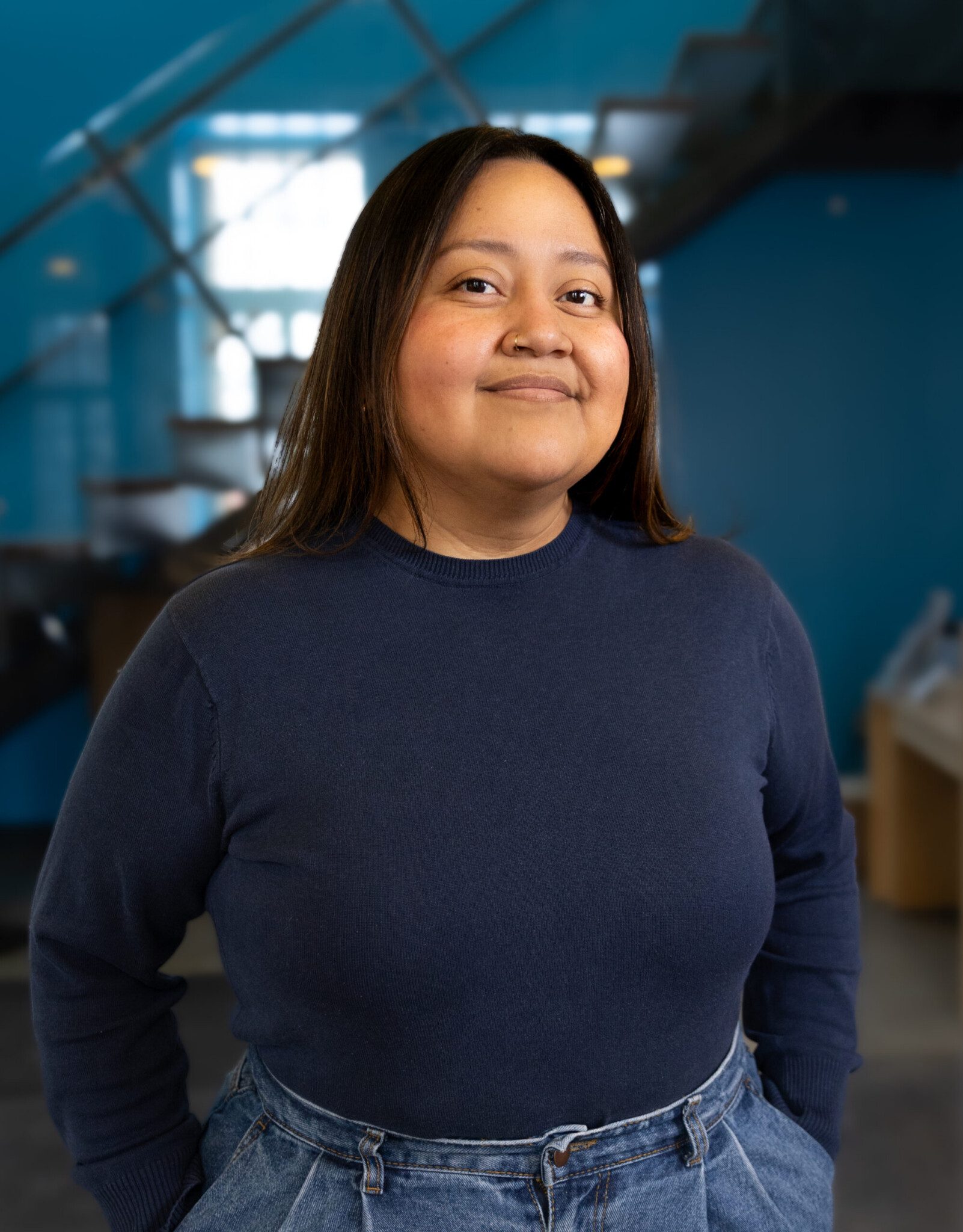 A person with long brown hair, wearing a navy blue sweater and blue jeans, stands indoors in front of a blurred staircase and blue wall, smiling slightly at the camera.