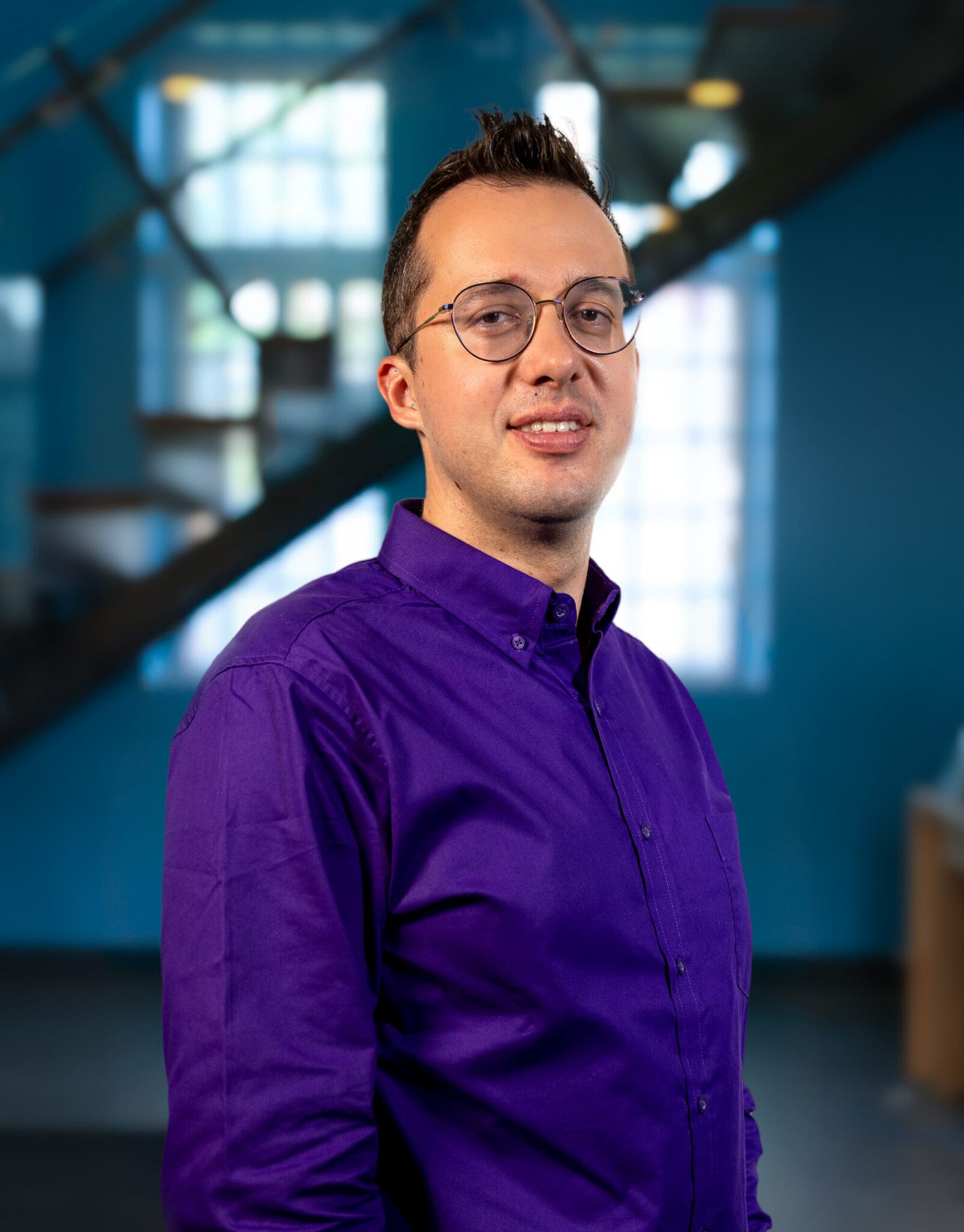A person wearing glasses and a purple button-up shirt stands indoors, smiling at the camera. The background is blurred, showing blue walls, large windows, and a staircase.