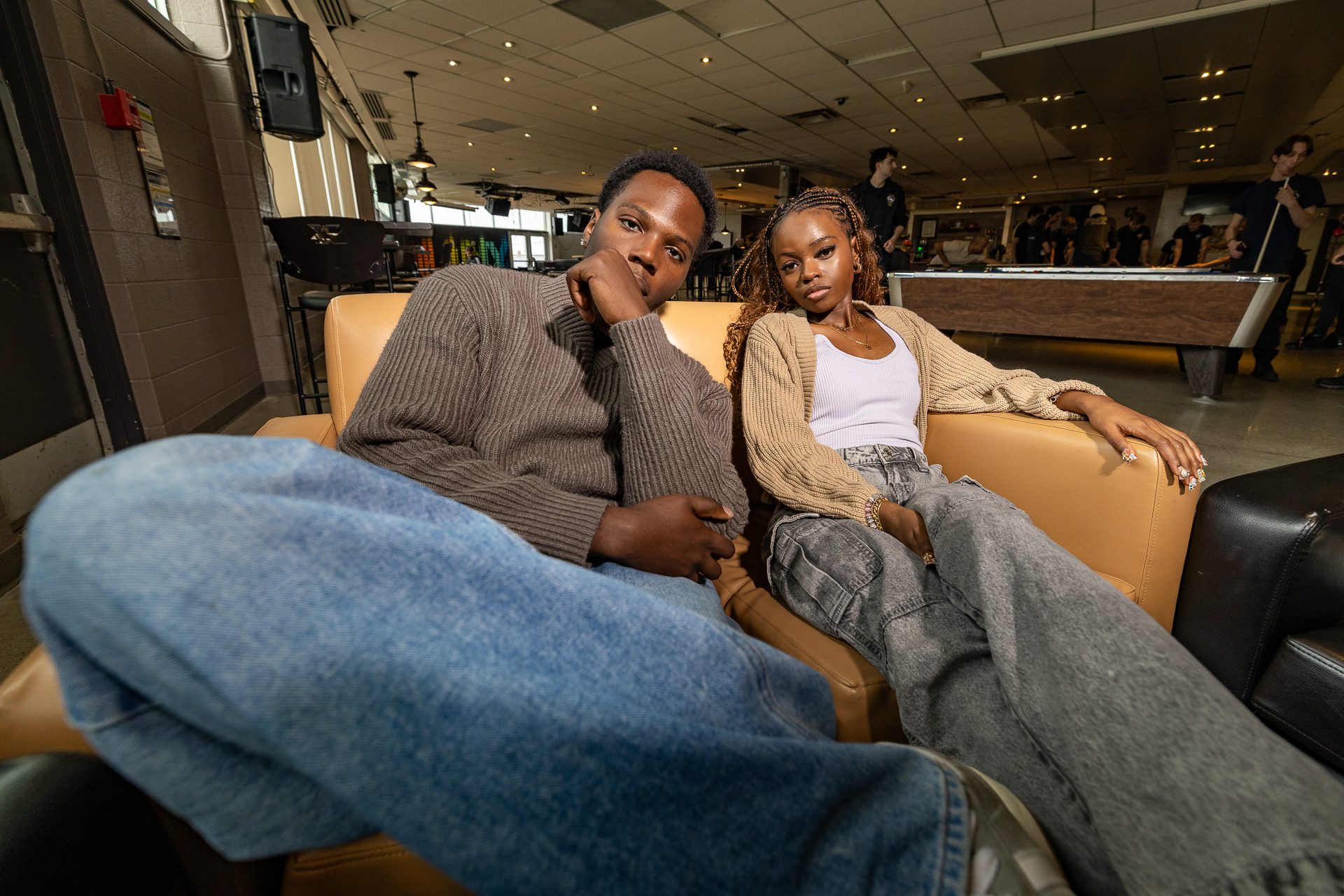 Two young adults lean on a tan leather couch in a busy indoor lounge, facing the camera.