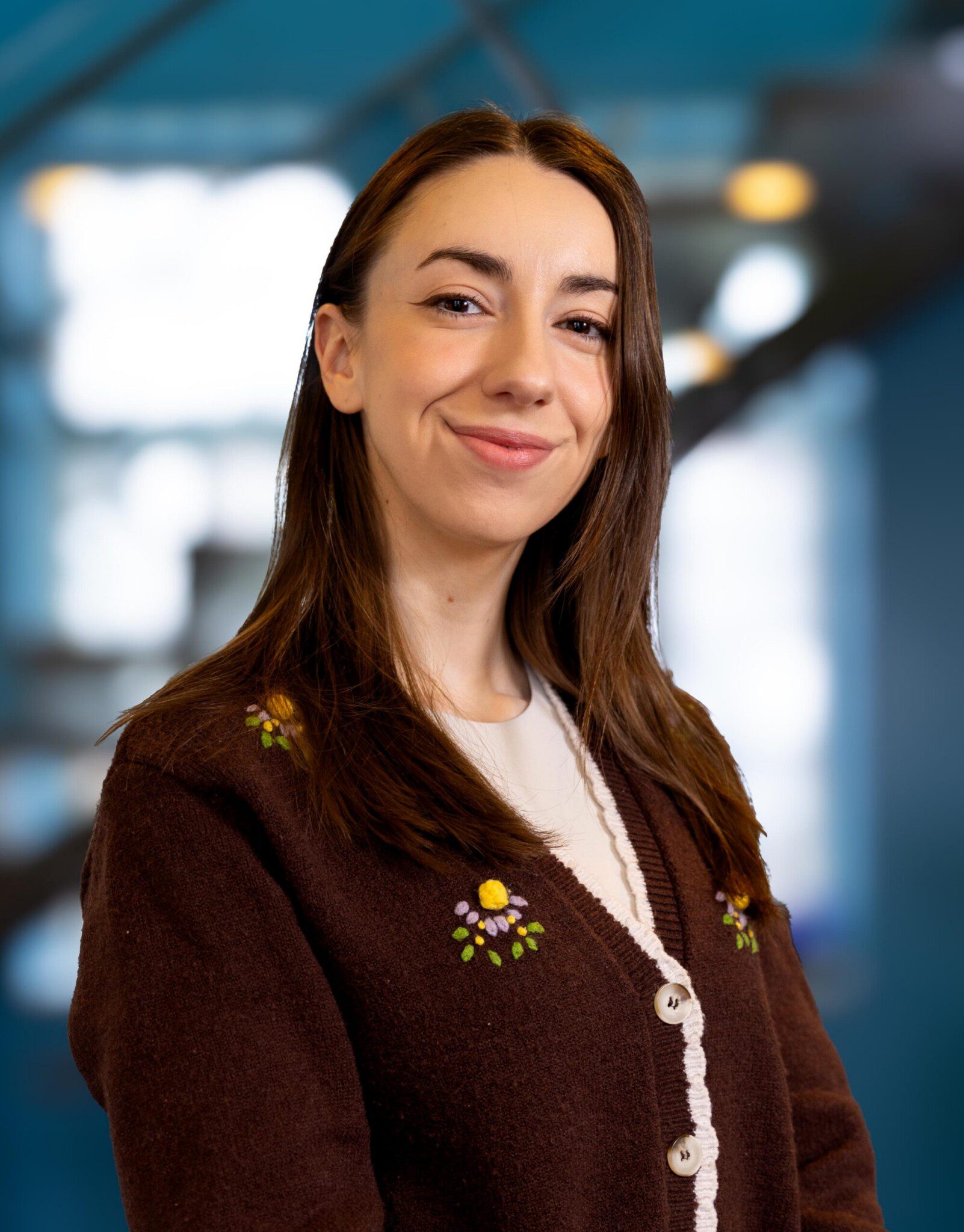 A young woman with long brown hair, wearing a brown sweater with floral embroidery over a white top, smiles confidently in a brightly lit indoor setting with a blue, blurred background.