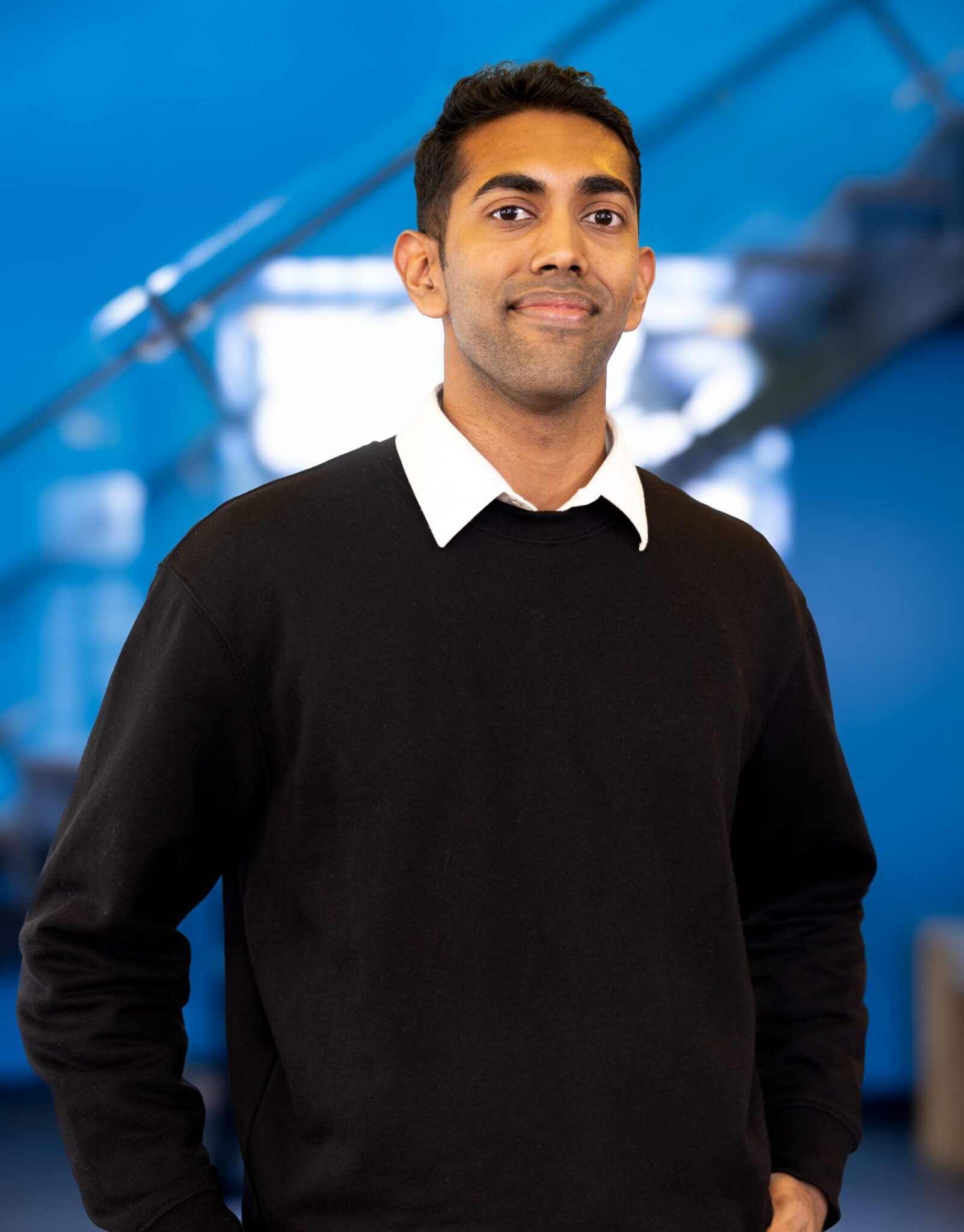 A person with short dark hair wearing a black sweater over a white collared shirt stands indoors, smiling slightly, with a blurred blue background and staircase behind them.