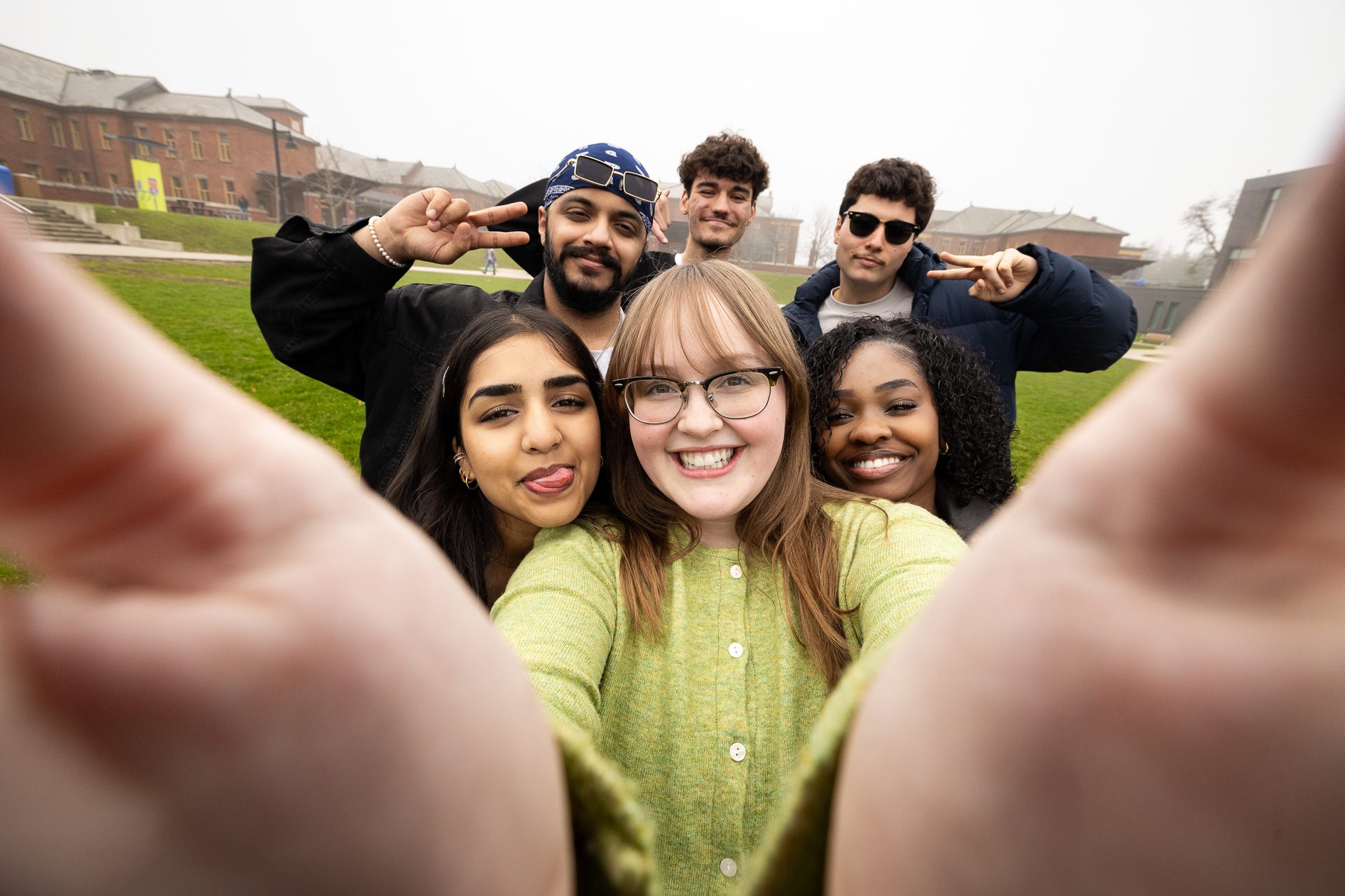 A group of six young adults stand close together outdoors, smiling and making peace signs as they pose for a cheerful selfie on a grassy area with buildings in the background.