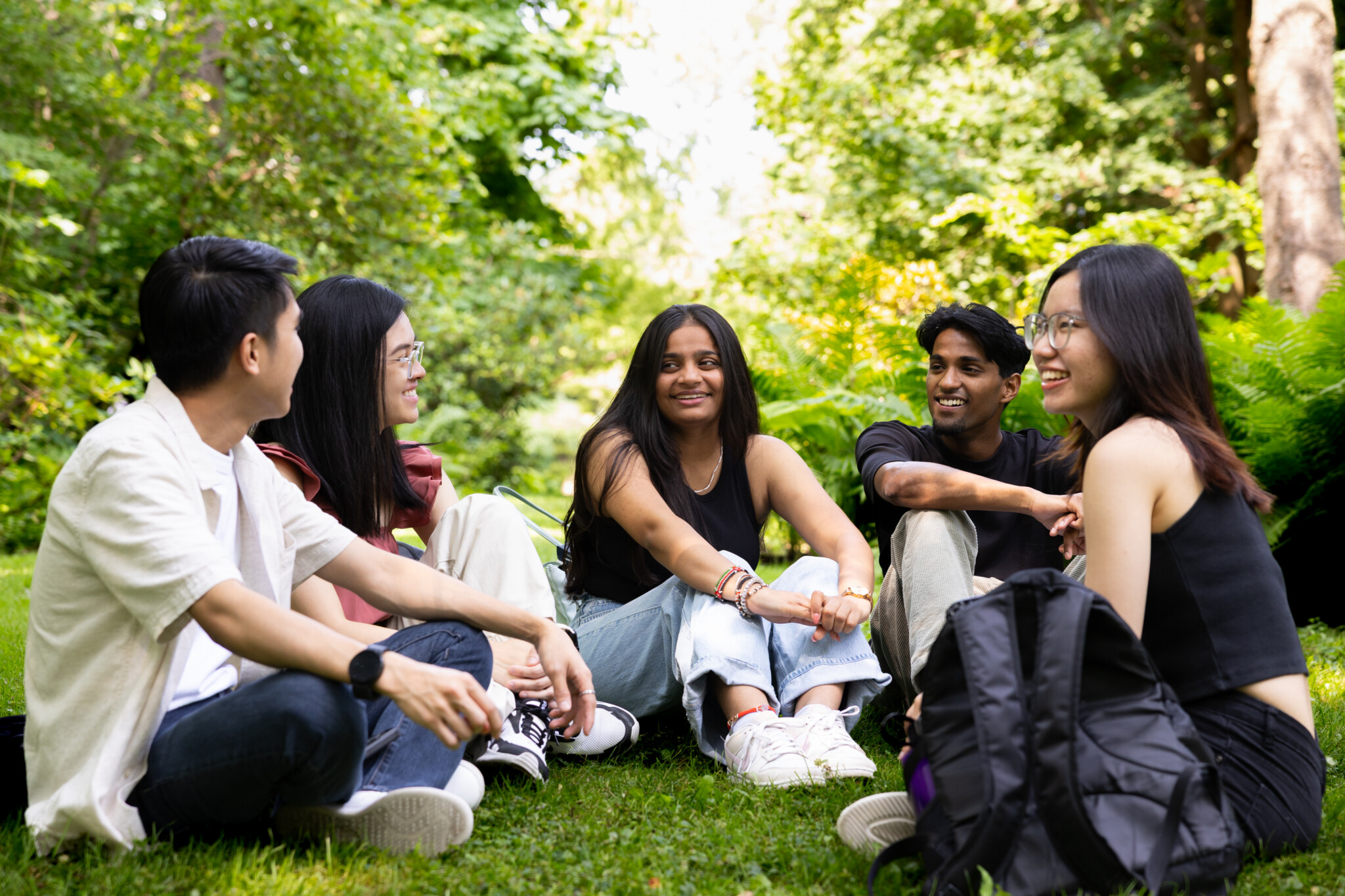 Five diverse students sitting casually in a circle on green grass in a lush outdoor setting, smiling and chatting with each other against a backdrop of trees and ferns, with a black backpack resting in the right foreground.