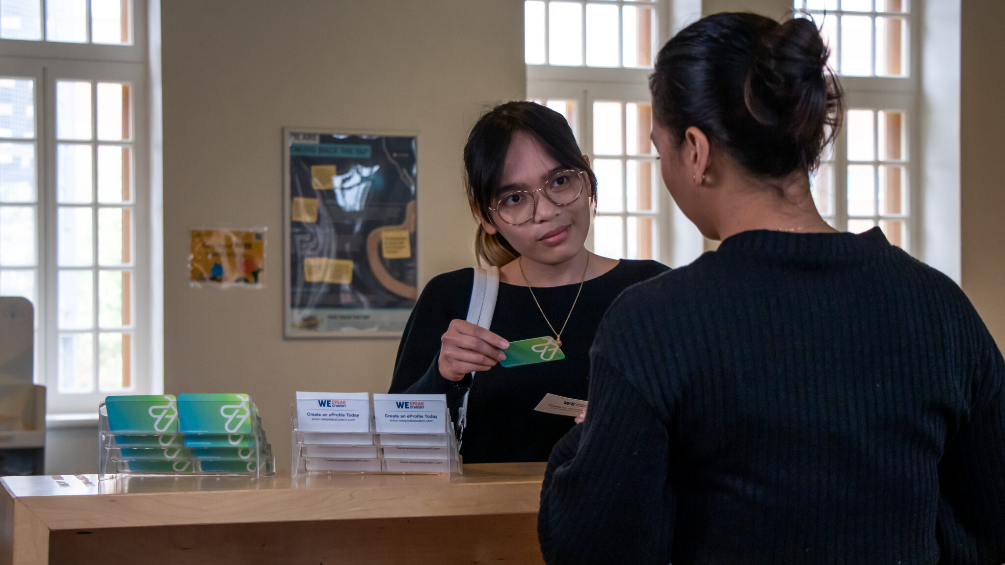 Two women stand at an indoor counter. One woman, wearing glasses, holds a card and faces the other woman. Brochures and green cards are displayed on the counter in a well-lit room with large windows.