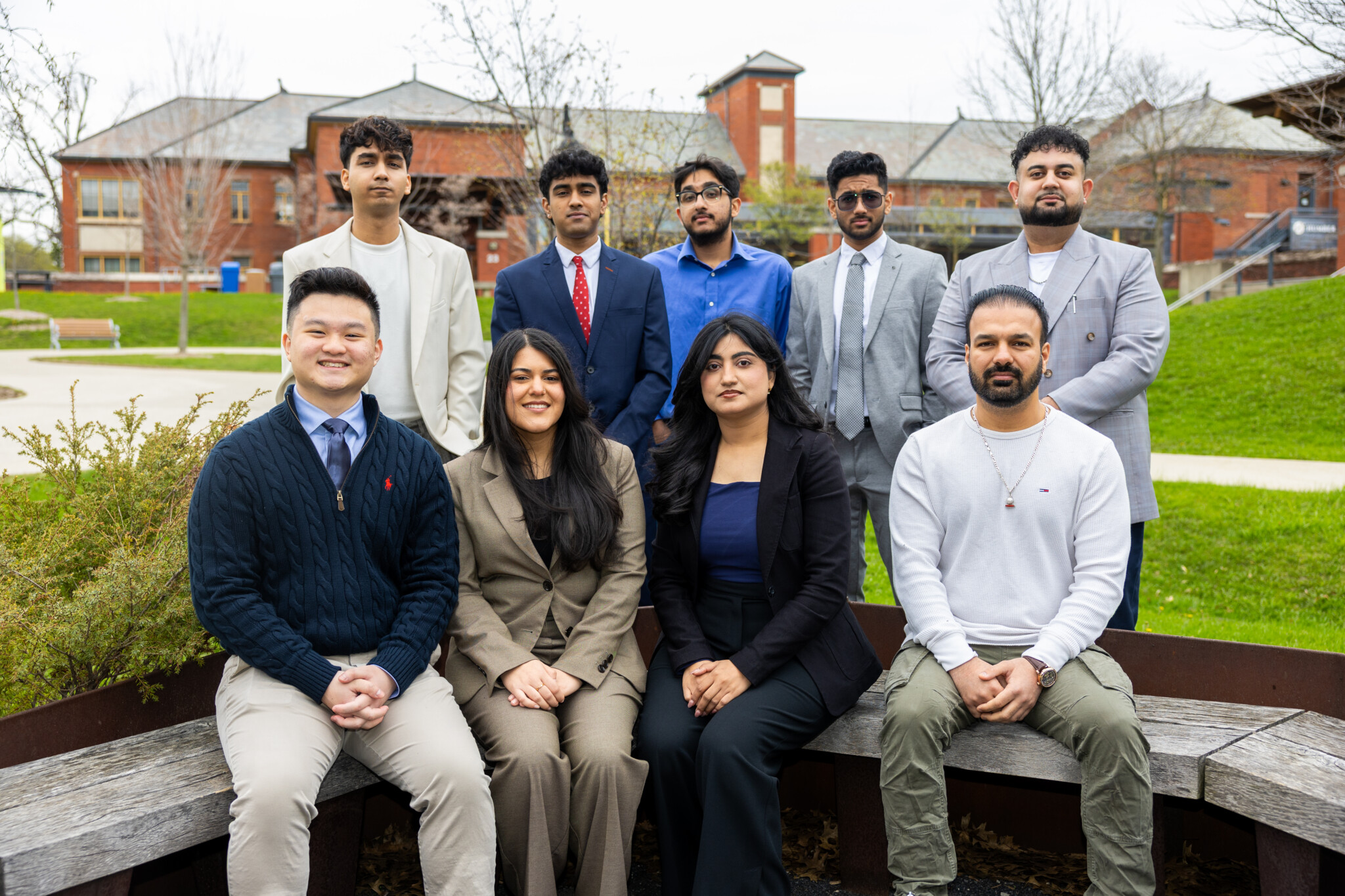 Nine people, dressed in business and casual attire, pose outdoors on a wooden bench and grass, with a brick building and trees in the background. The group looks toward the camera, smiling or maintaining neutral expressions.