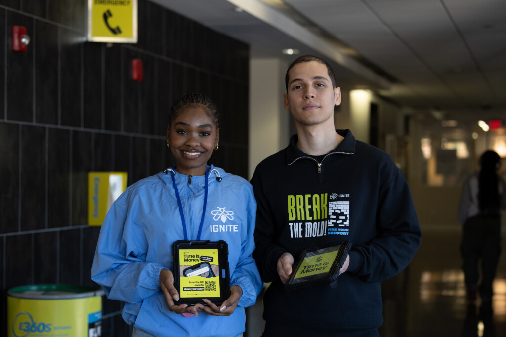 Two young adults stand indoors, both holding boxes labeled “Time for kindness” with QR codes. The woman wears a blue “IGNITE” jacket and smiles; the man wears a black “Break the Mould” shirt. They stand in a hallway with signs and a trash bin.
