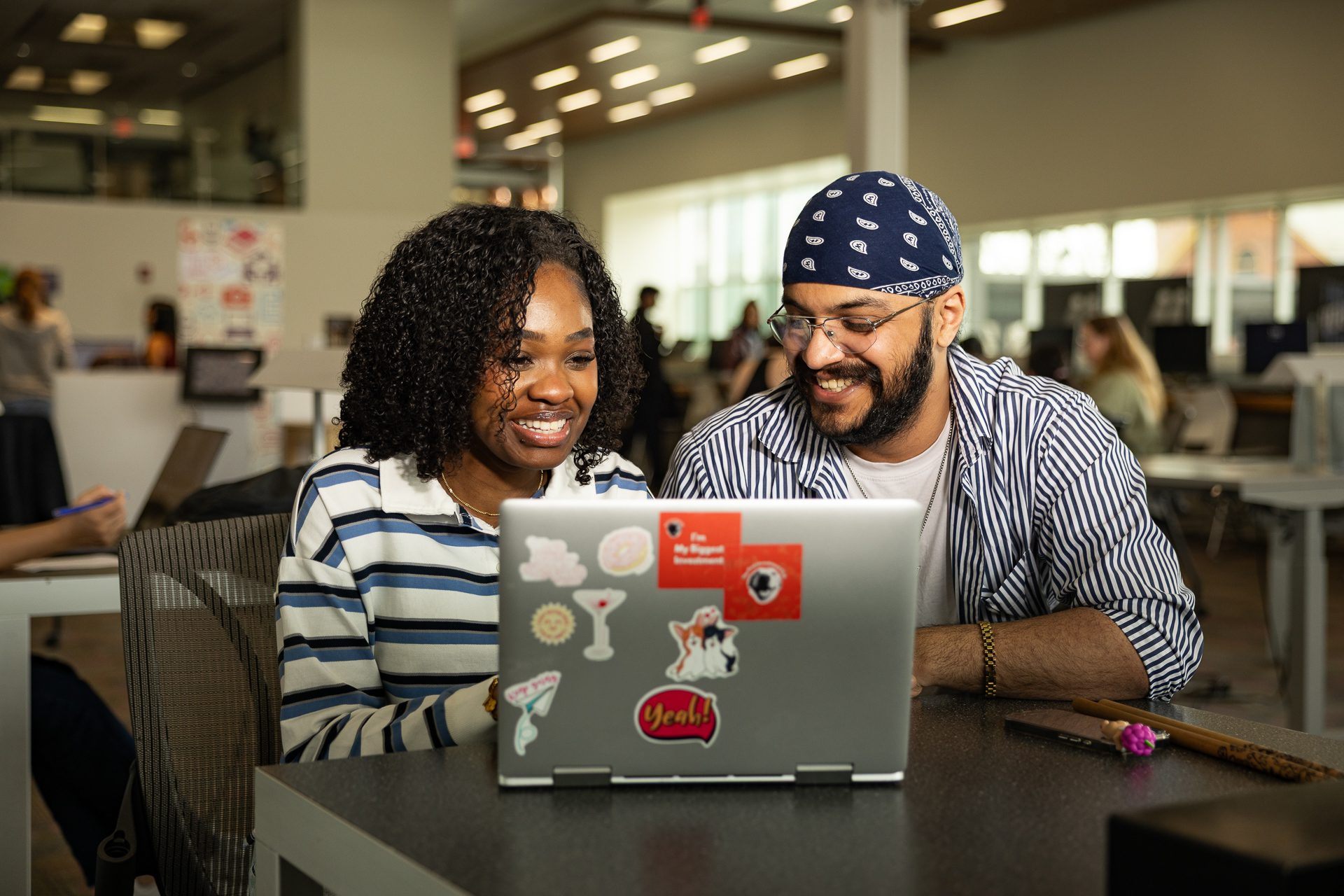 Two coworkers smile while looking at a sticker-covered laptop in a bright open office.