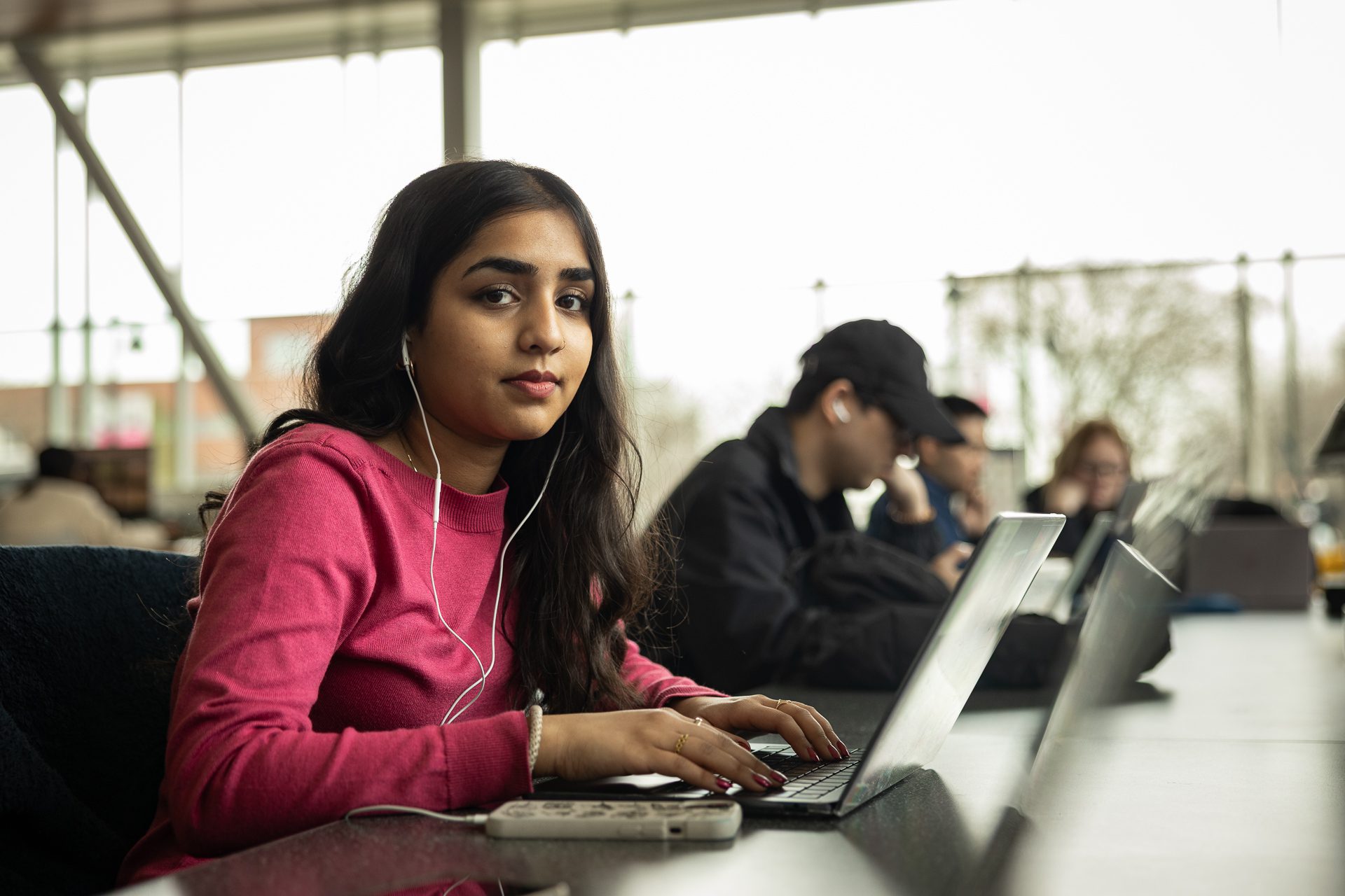 A young woman with long dark hair, wearing a pink sweater and earphones, sits at a desk using a laptop in a bright, modern study area with other people working in the background.