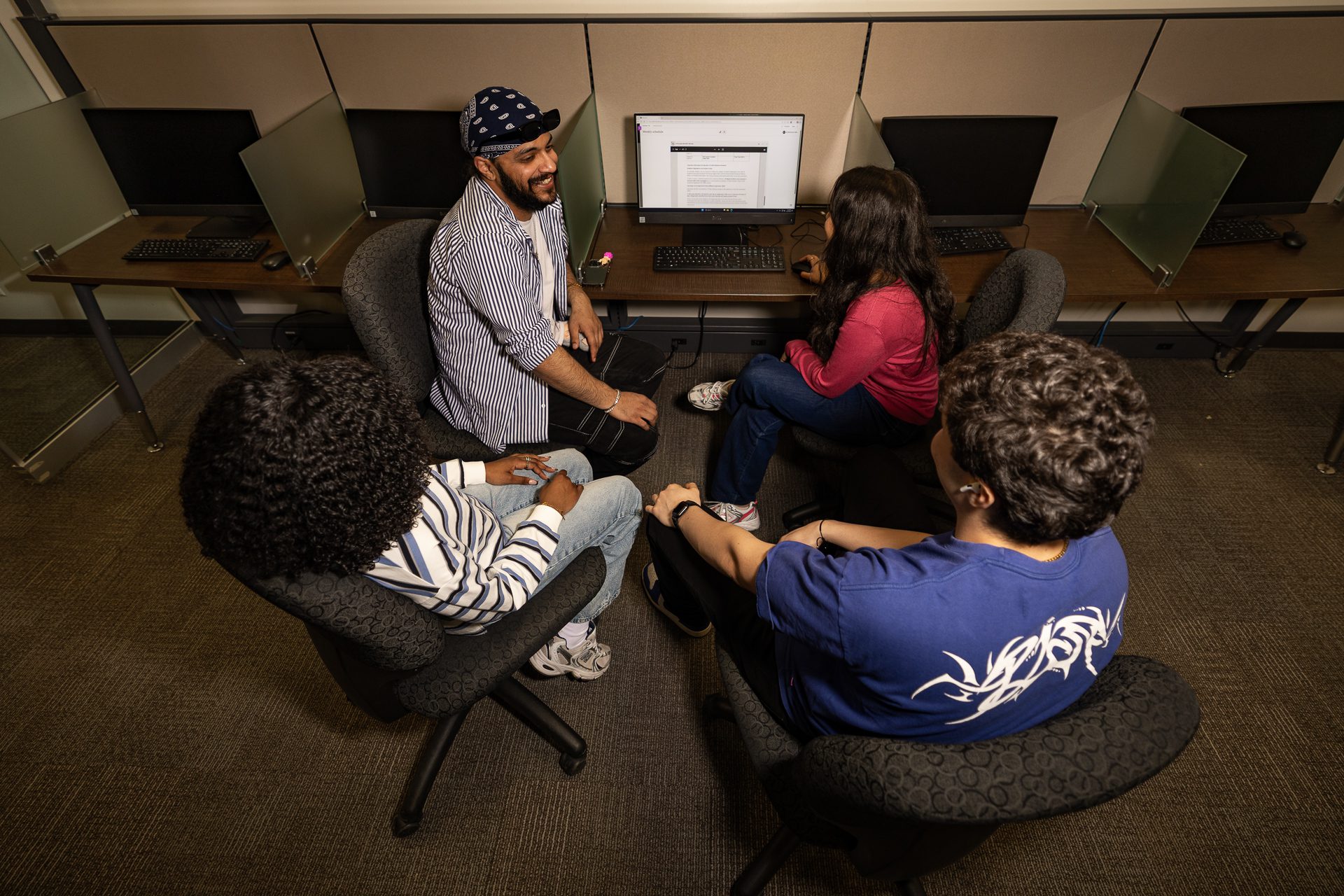Group of students in a computer lab sitting in a circle around a shared desk, smiling and chatting while one person uses the computer.