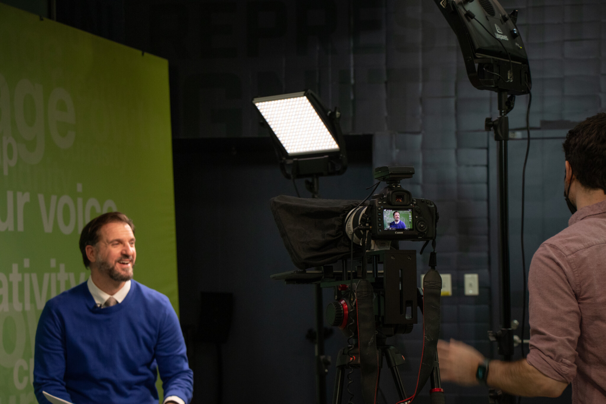 A man in a blue sweater sits smiling in front of a green backdrop during a video shoot, while another person stands near a camera and studio lights.