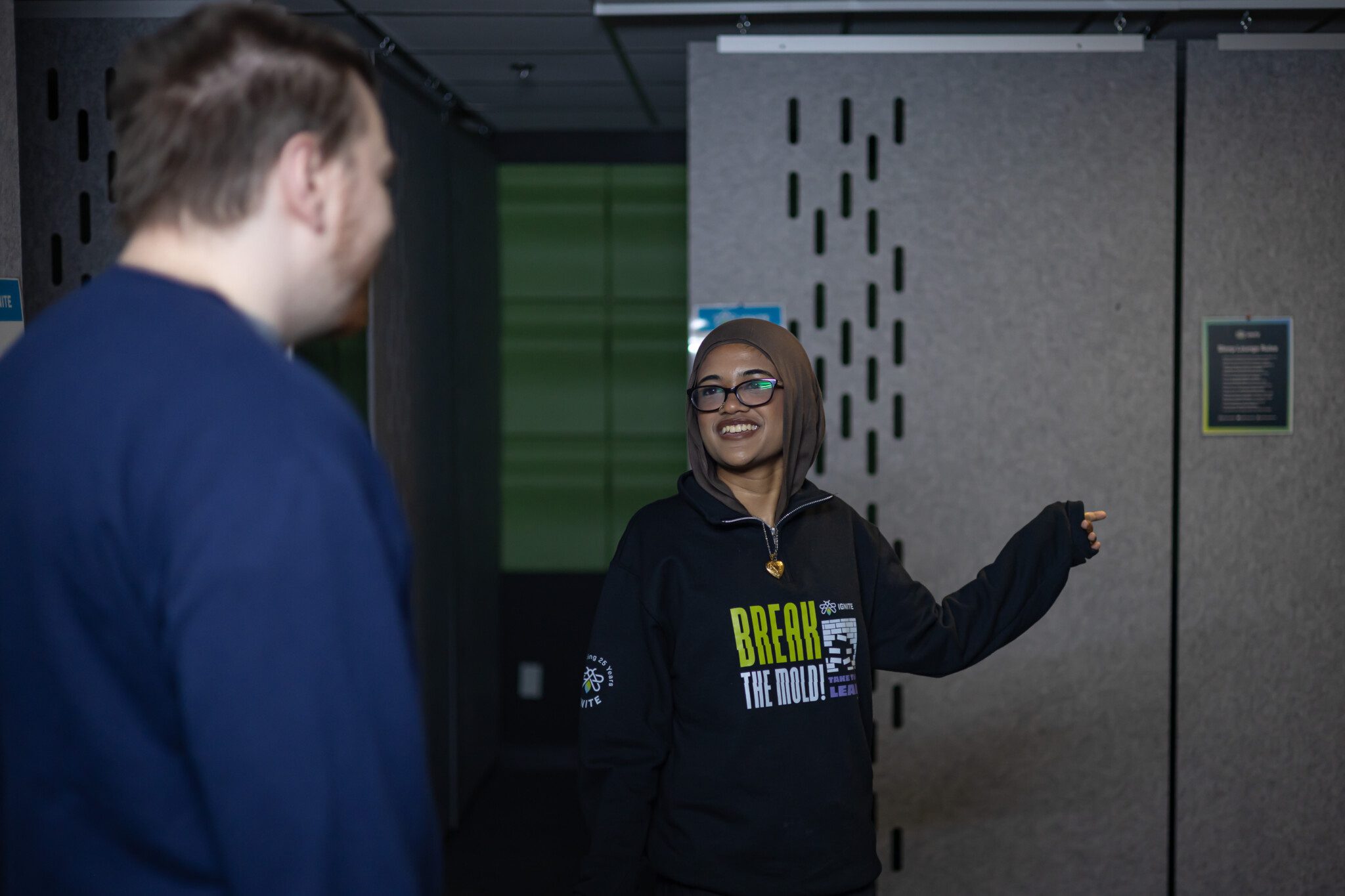 A female IGNITE staff member wearing a brown hijab, glasses, and a black "BREAK THE MOLD!" sweatshirt smiles as she gestures with her hand toward a sleep pod room while assisting a male student in a blue sweater.