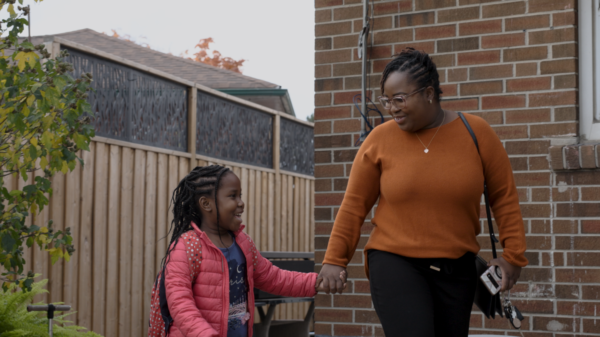A woman in an orange sweater and glasses smiles while holding hands with a young girl in a pink jacket and backpack as they walk outside near a brick building and a wooden fence.