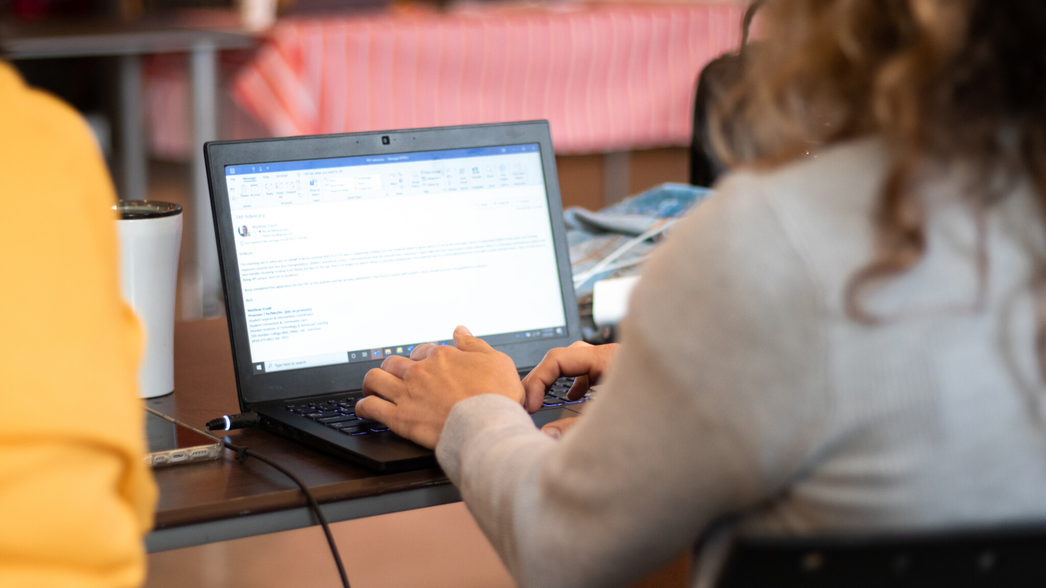 A person with curly hair types on a laptop at a table, with an email or document open on the screen. A coffee cup and some other items are on the desk nearby. The background is softly blurred.