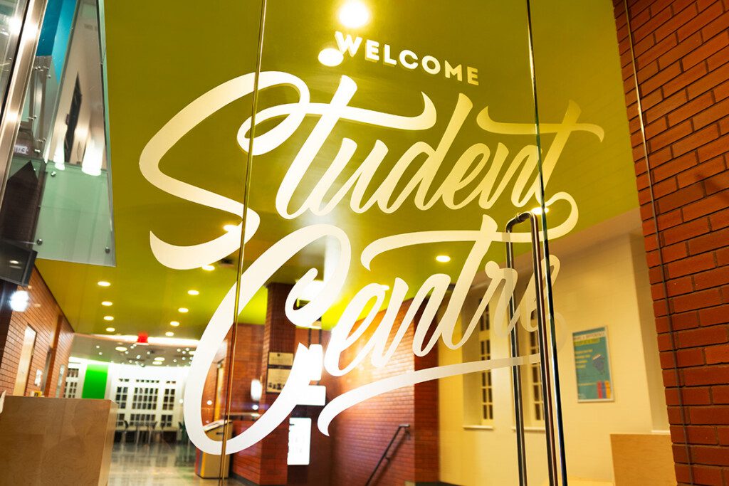 Glass doors with large white script text reading Welcome Student Centre. Brightly lit interior with a green ceiling, brick walls, and a colorful poster on the wall inside.