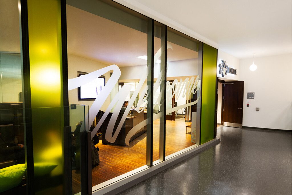 Glass office with large frosted Welcome lettering on the window, showing a modern interior with wooden floors and seating. The hallway outside has a sign for Suite and a wall-mounted light.