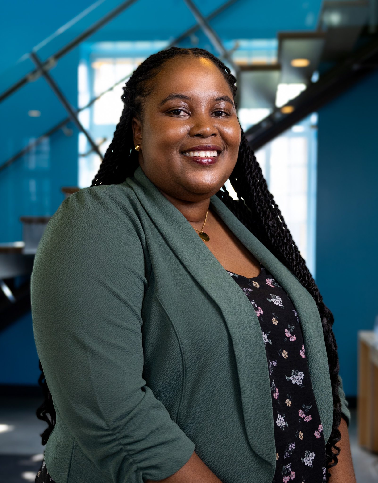 A woman with long braided hair, wearing a green blazer over a floral top, smiles while posing indoors in front of a blue wall and a staircase.