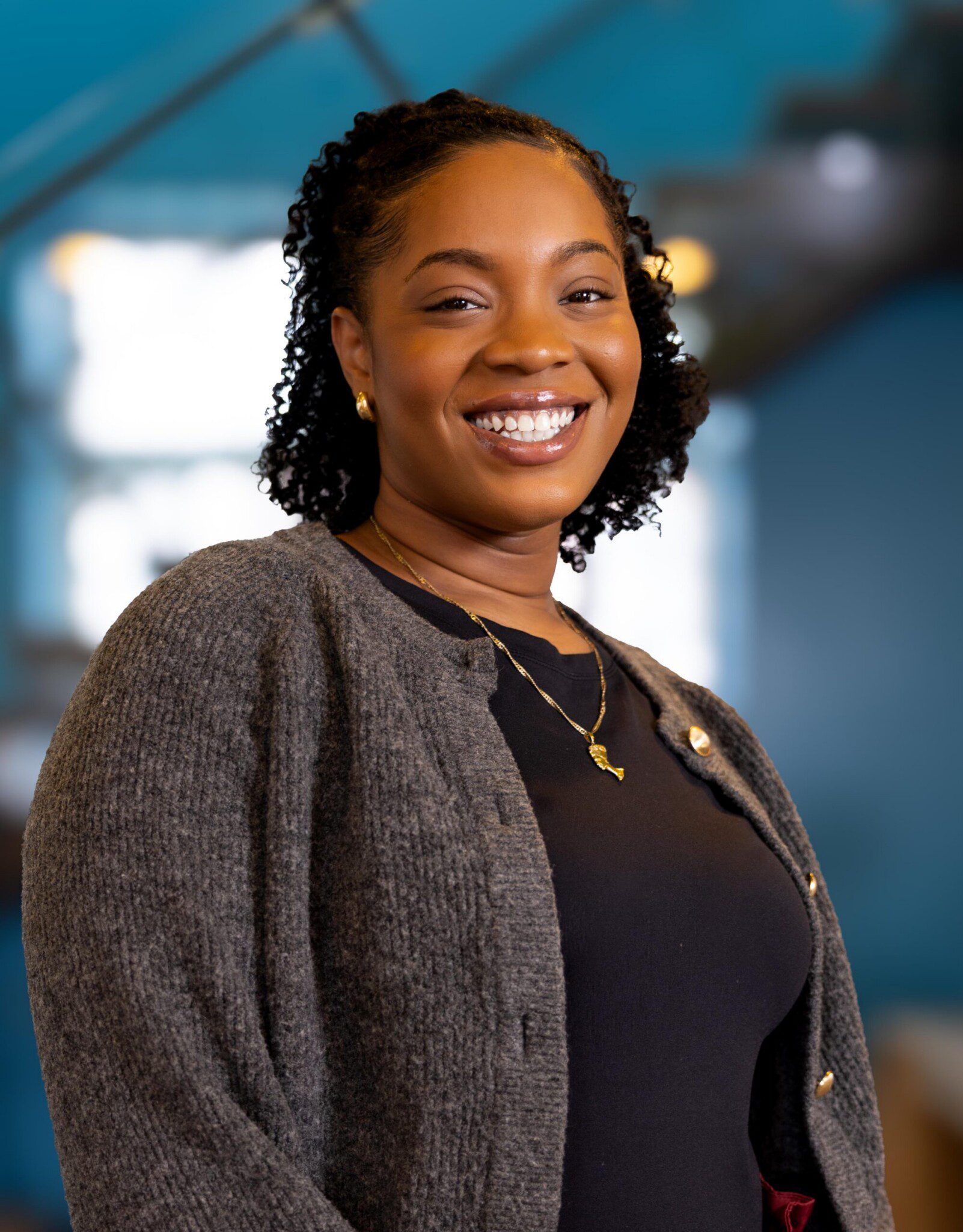 A smiling woman with curly hair, wearing a dark top, gray cardigan, and gold necklace, stands indoors with a blurred blue background and soft lighting.