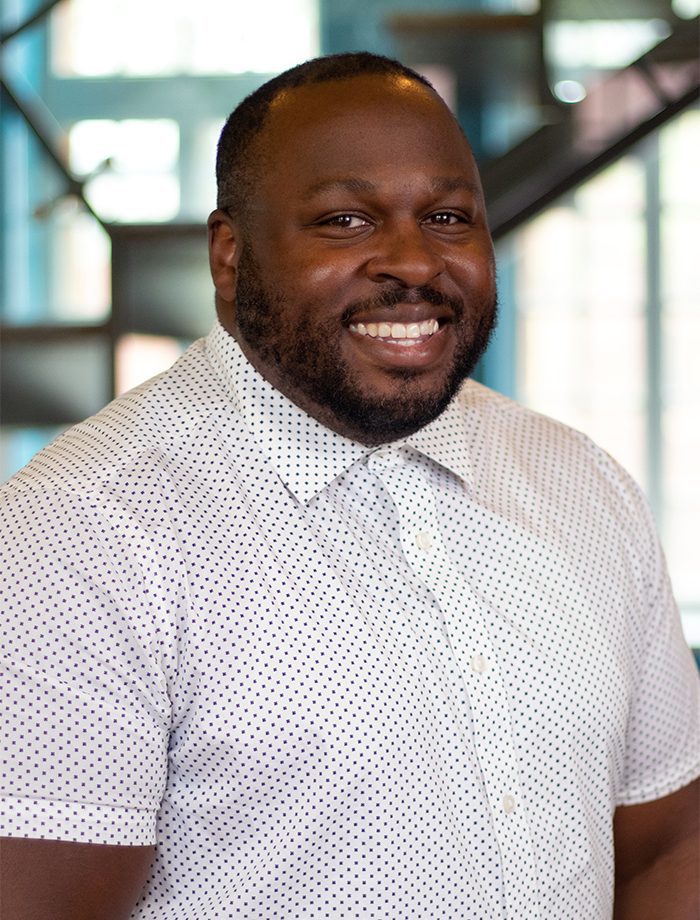 A smiling man with a beard, wearing a white short-sleeve button-up shirt with small black dots, stands indoors in front of a blurred modern background with windows and stairs.