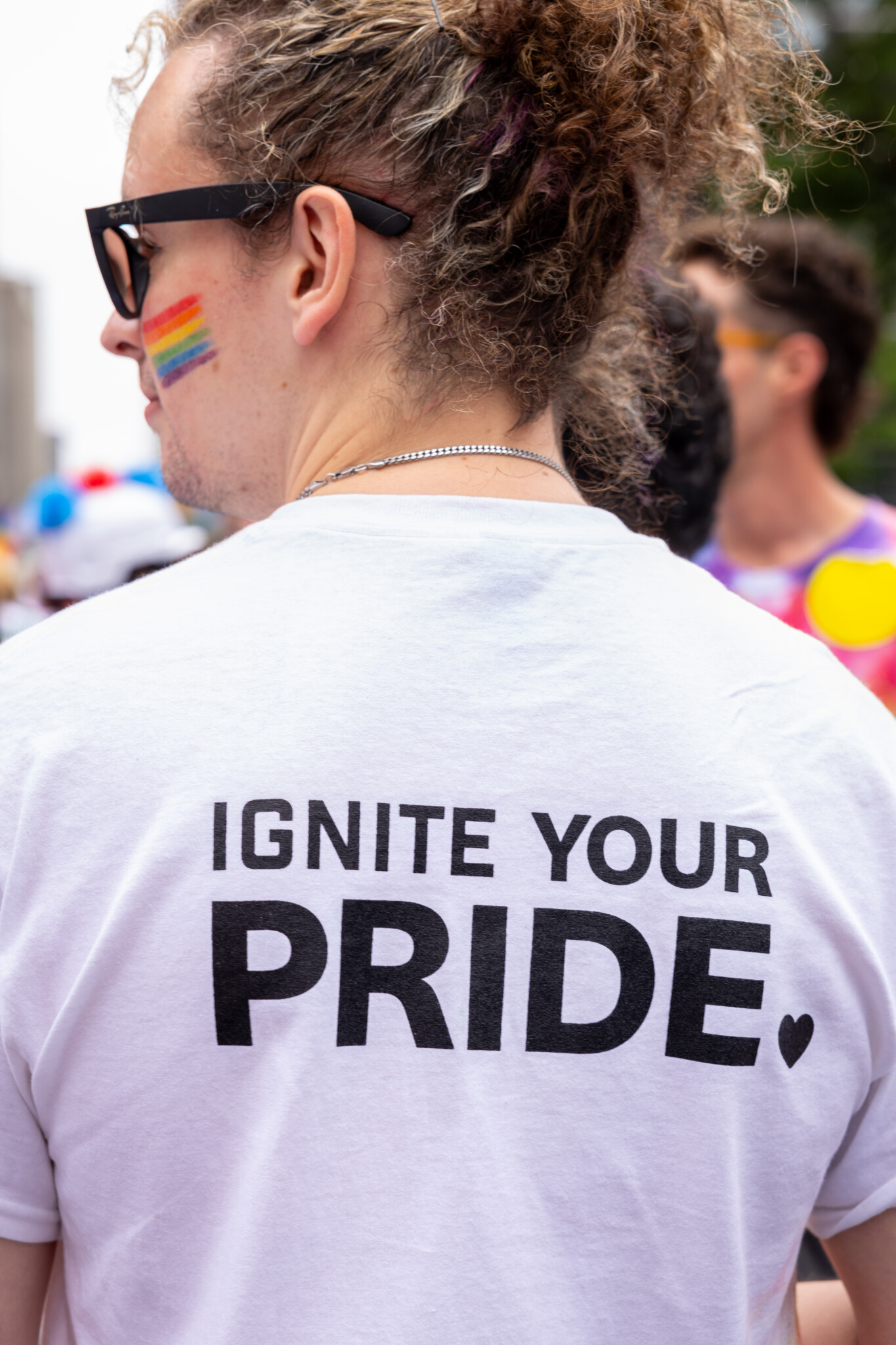 A person with curly hair, rainbow face paint, and a white shirt reading IGNITE YOUR PRIDE stands outdoors at a pride event.