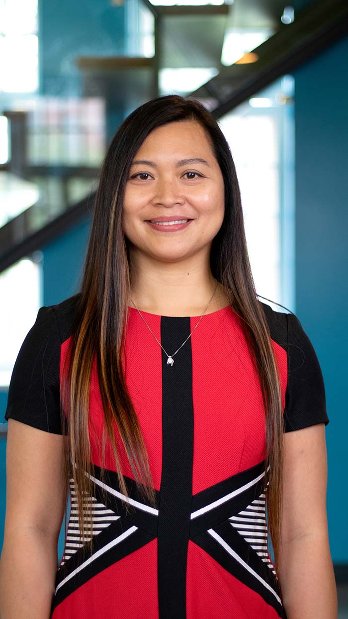A woman with long straight hair wearing a red and black dress stands smiling in front of a blue wall and a staircase.