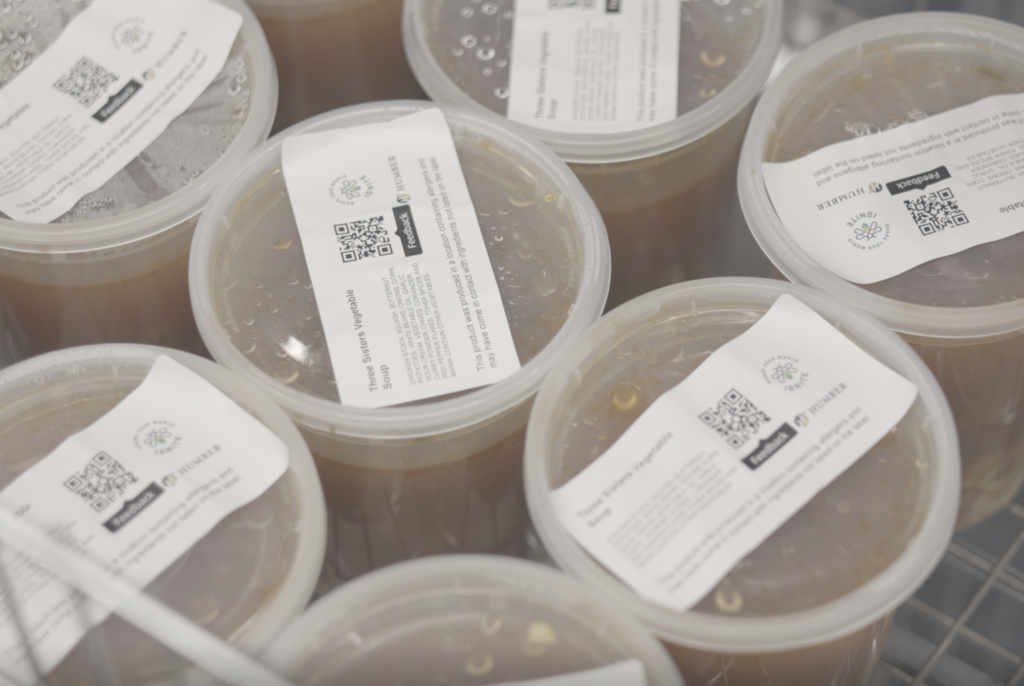 Several clear plastic containers filled with brown liquid, each sealed with a lid and labeled with printed white stickers containing QR codes and text, arranged closely together in a refrigerated setting.