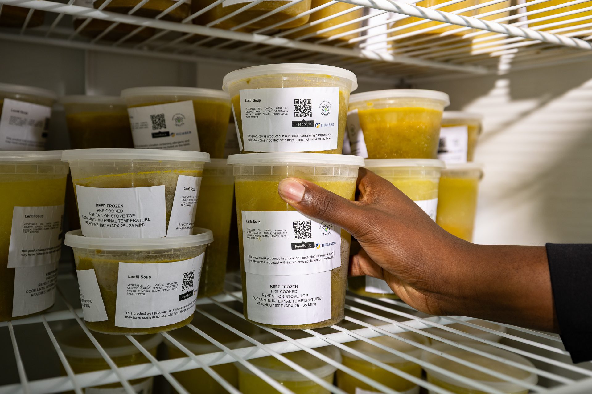 A hand reaches for a plastic container of lentil soup among several similar containers with printed labels, all stored on a white wire refrigerator shelf.
