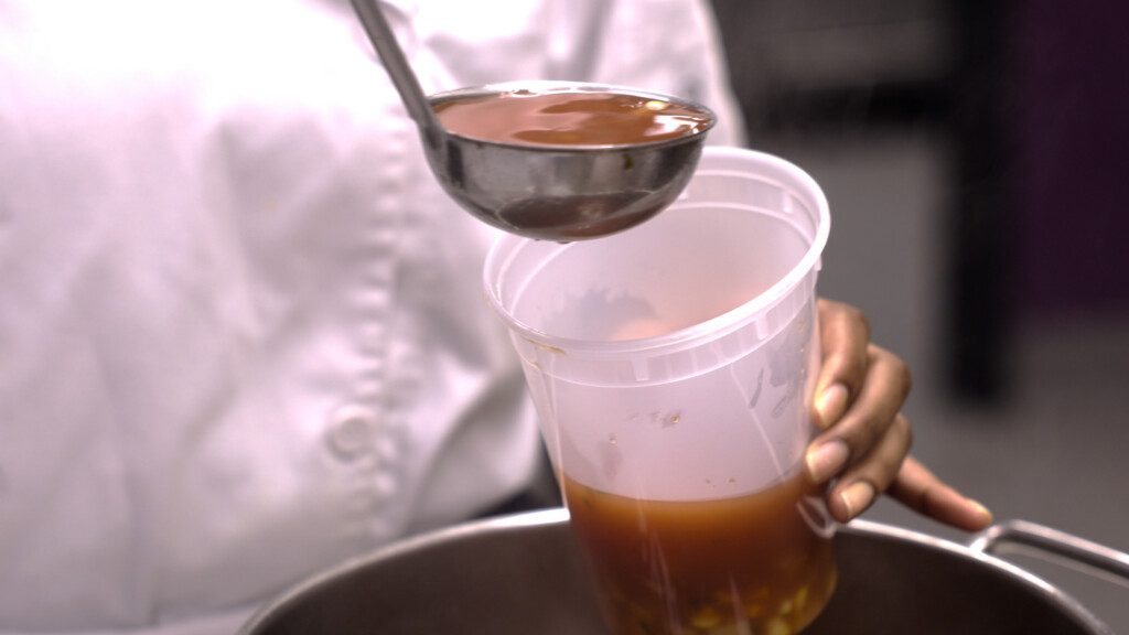 A person in a white chef coat pours reddish-brown soup with chunks of vegetables from a ladle into a clear plastic container.