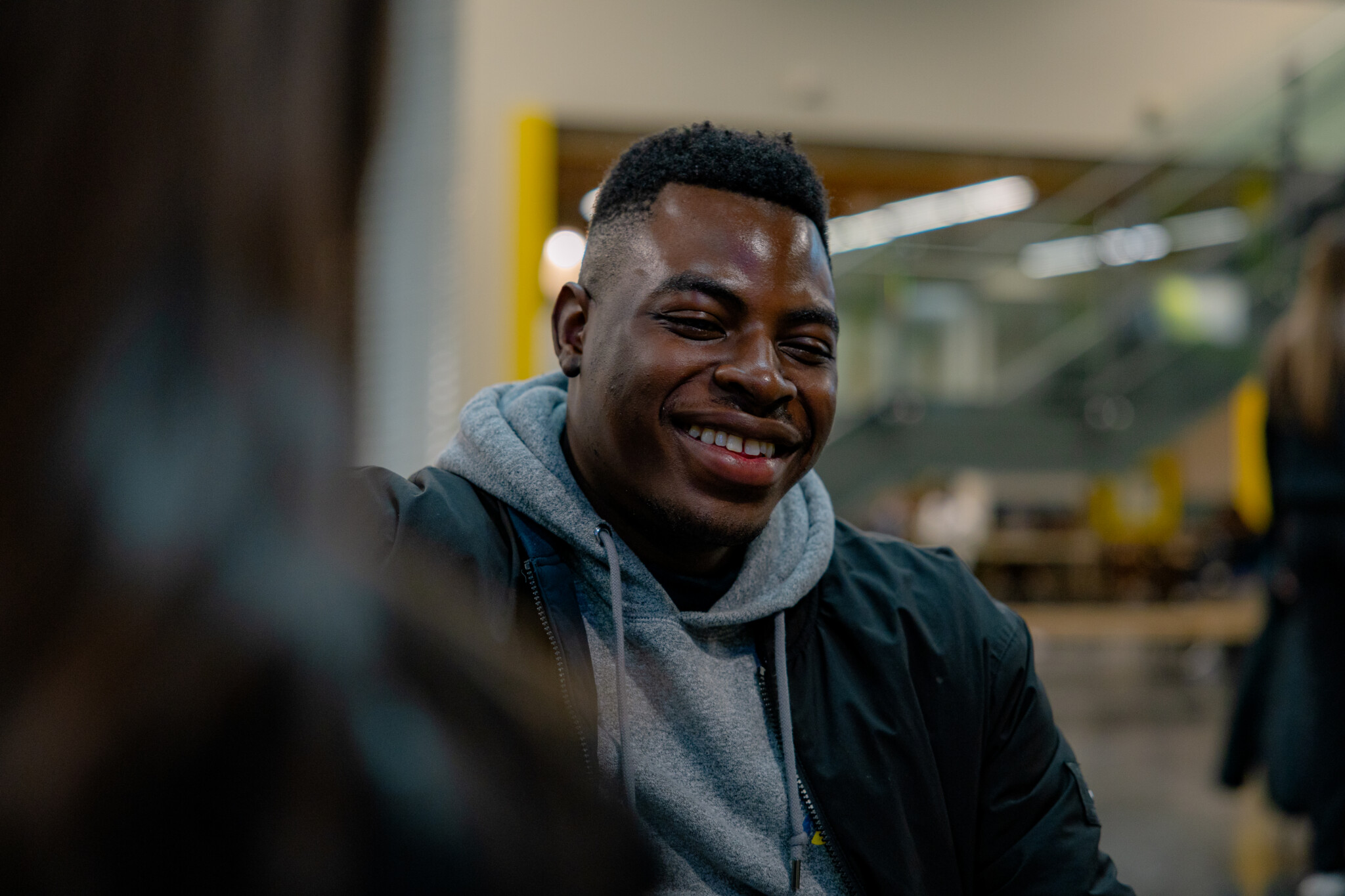 A man wearing a black jacket and gray hoodie smiles warmly while sitting indoors in a modern, brightly lit space. The background is slightly blurred, and another person is visible in the foreground.
