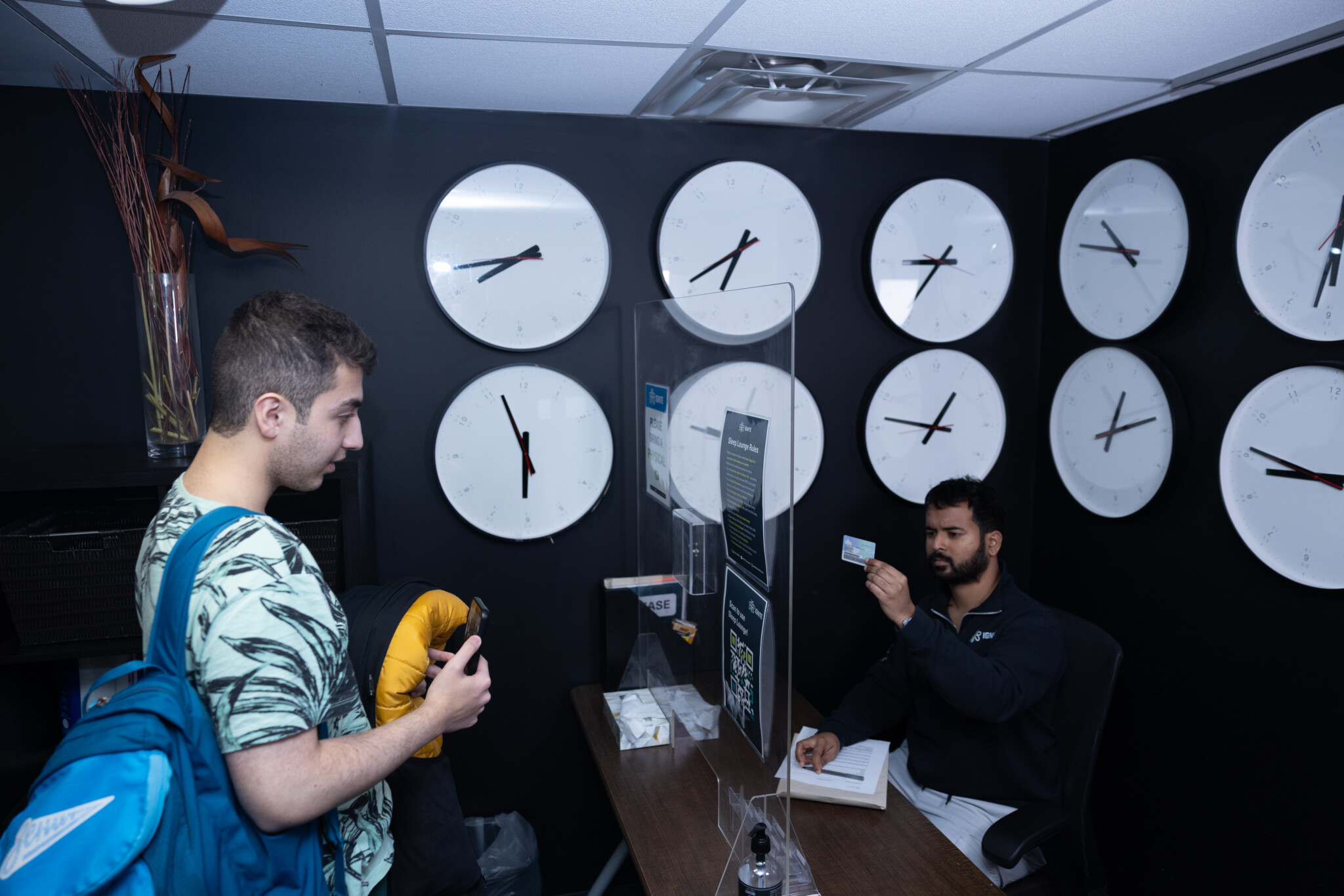 A male student carrying a blue backpack stands at a reception desk presenting his student ID to an IGNITE staff member, who holds up the card to verify it, inside a room with dark walls decorated with numerous large white wall clocks.