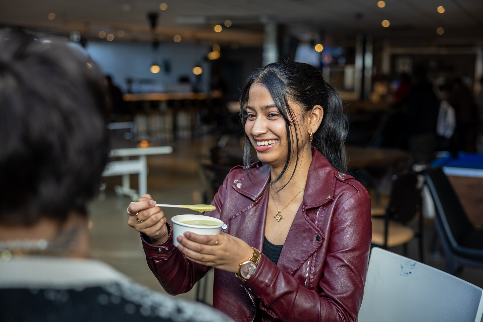 A smiling woman in a red leather jacket holds a cup and lifts a spoonful of soup, sitting indoors at a café or restaurant with a blurred background and another person in the foreground.