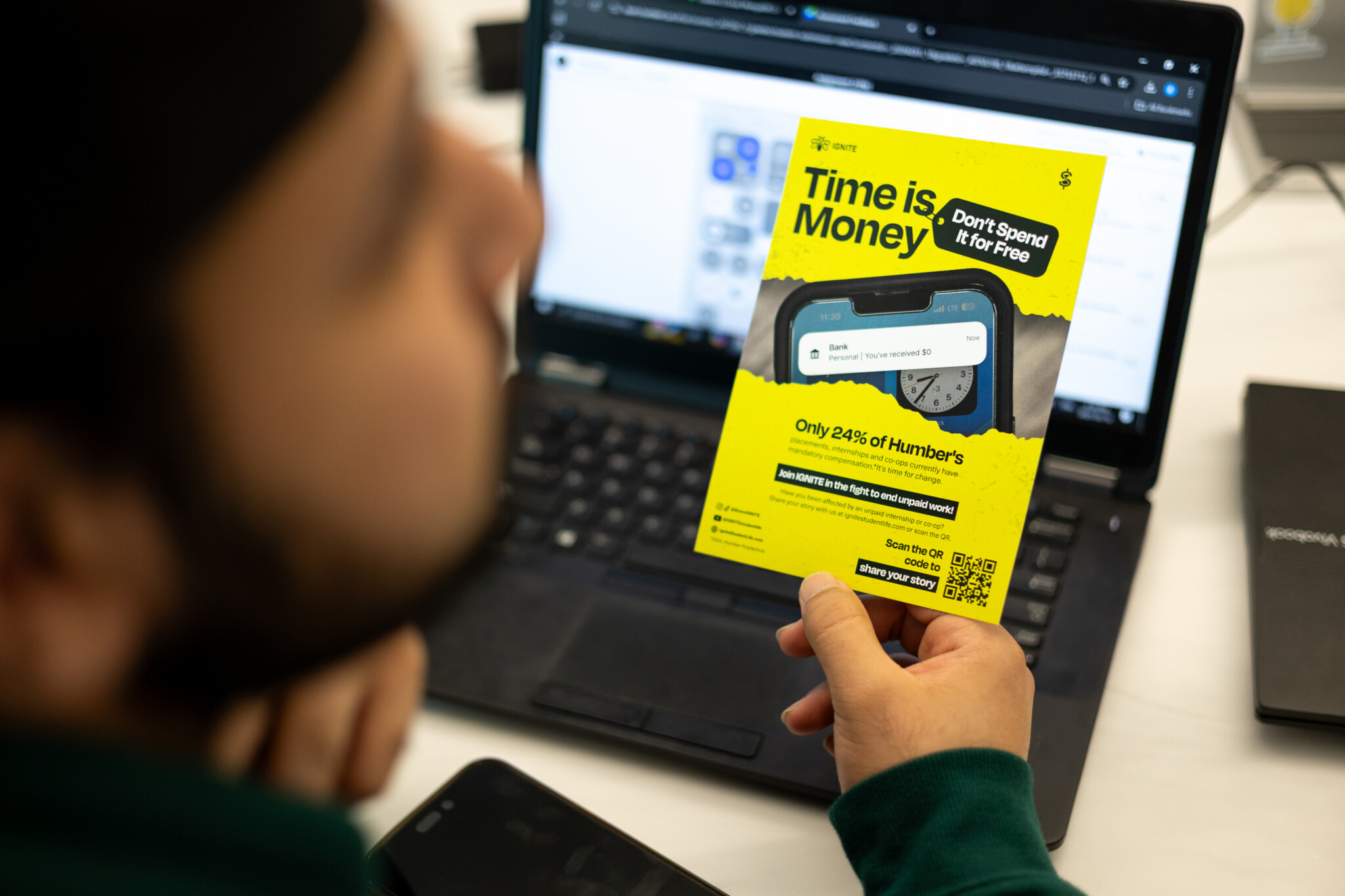 A person holds a yellow flyer reading Time is Money. Don’t Spend It for Free, in front of a laptop. The flyer displays a smartwatch and statistics, promoting efficiency and use of time.