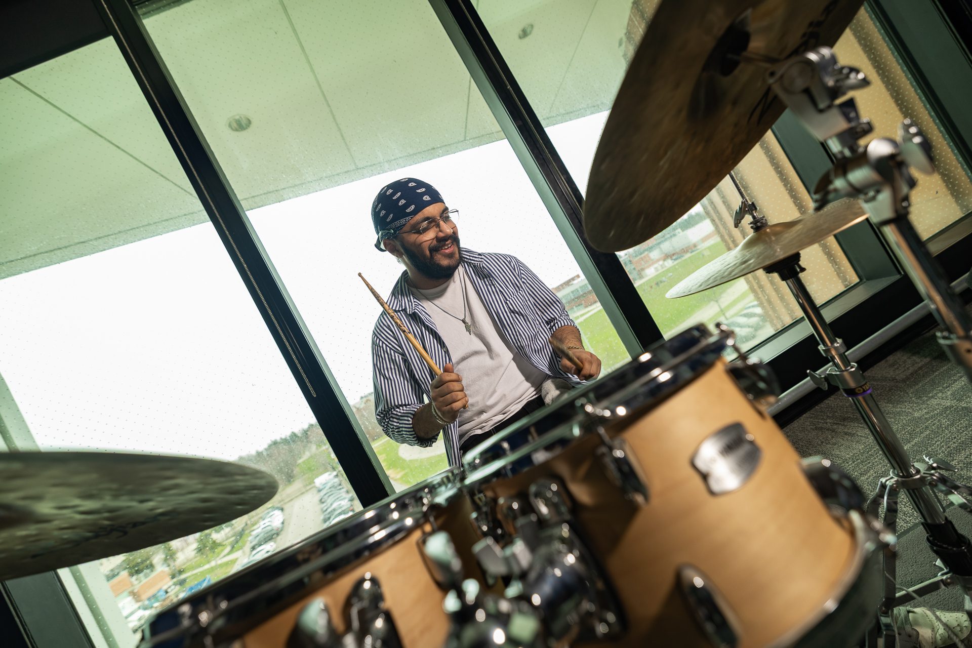 A smiling male student wearing a blue patterned bandana, glasses, and a striped button-down shirt holding drumsticks while playing a Yamaha acoustic drum set next to large windows overlooking campus grounds.