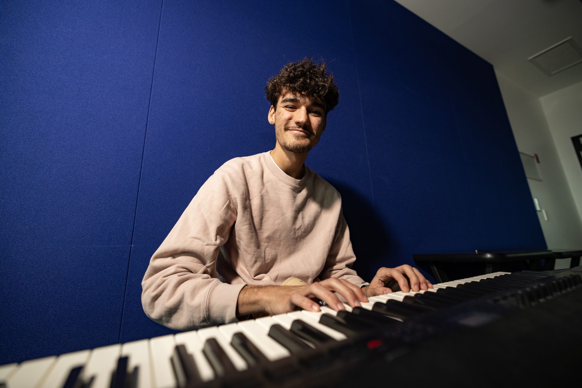 Low-angle shot of a smiling male student with curly hair wearing a light pink sweatshirt playing an electronic keyboard set up against a bright blue studio wall.