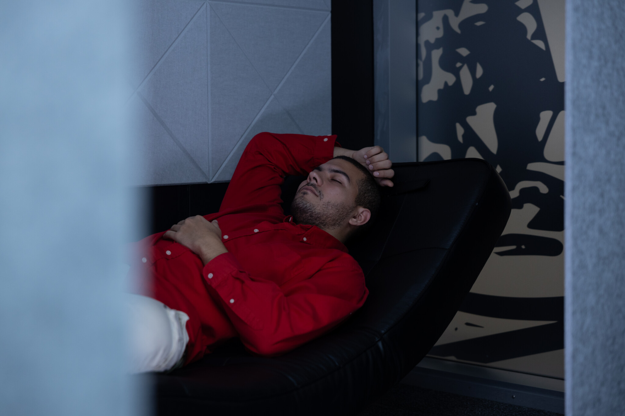 A young man in a red shirt taking a nap on a black curved lounge chair in a quiet, dark room.
