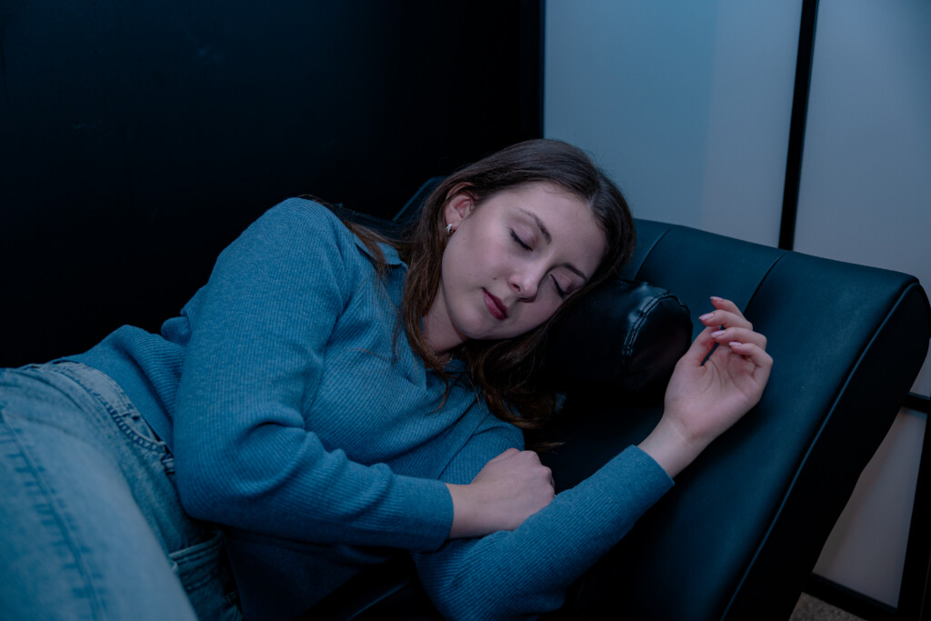 A young woman with long brown hair wearing a blue long-sleeved sweater lies peacefully asleep on a black leather lounge chair inside a dimly lit rest area.