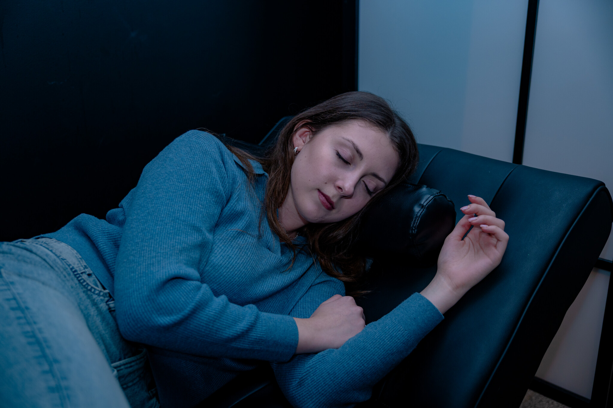 A young woman with long brown hair wearing a blue long-sleeved sweater lies peacefully asleep on a black leather lounge chair inside a dimly lit rest area.