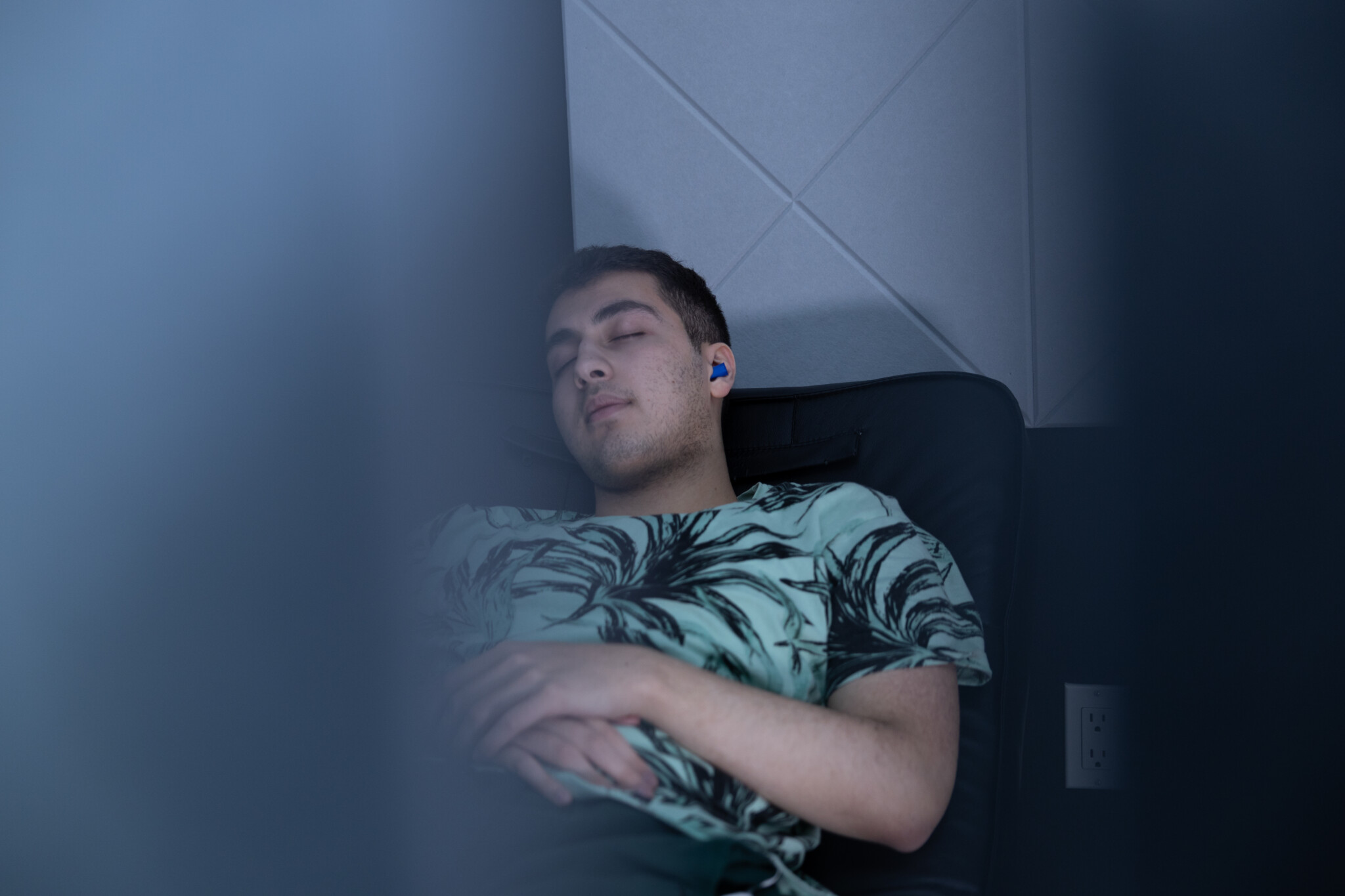 Close-up shot of a male student wearing a patterned green t-shirt and blue earplugs resting peacefully with his eyes closed in a dark, quiet sleep pod at the IGNITE Sleep Lounge.