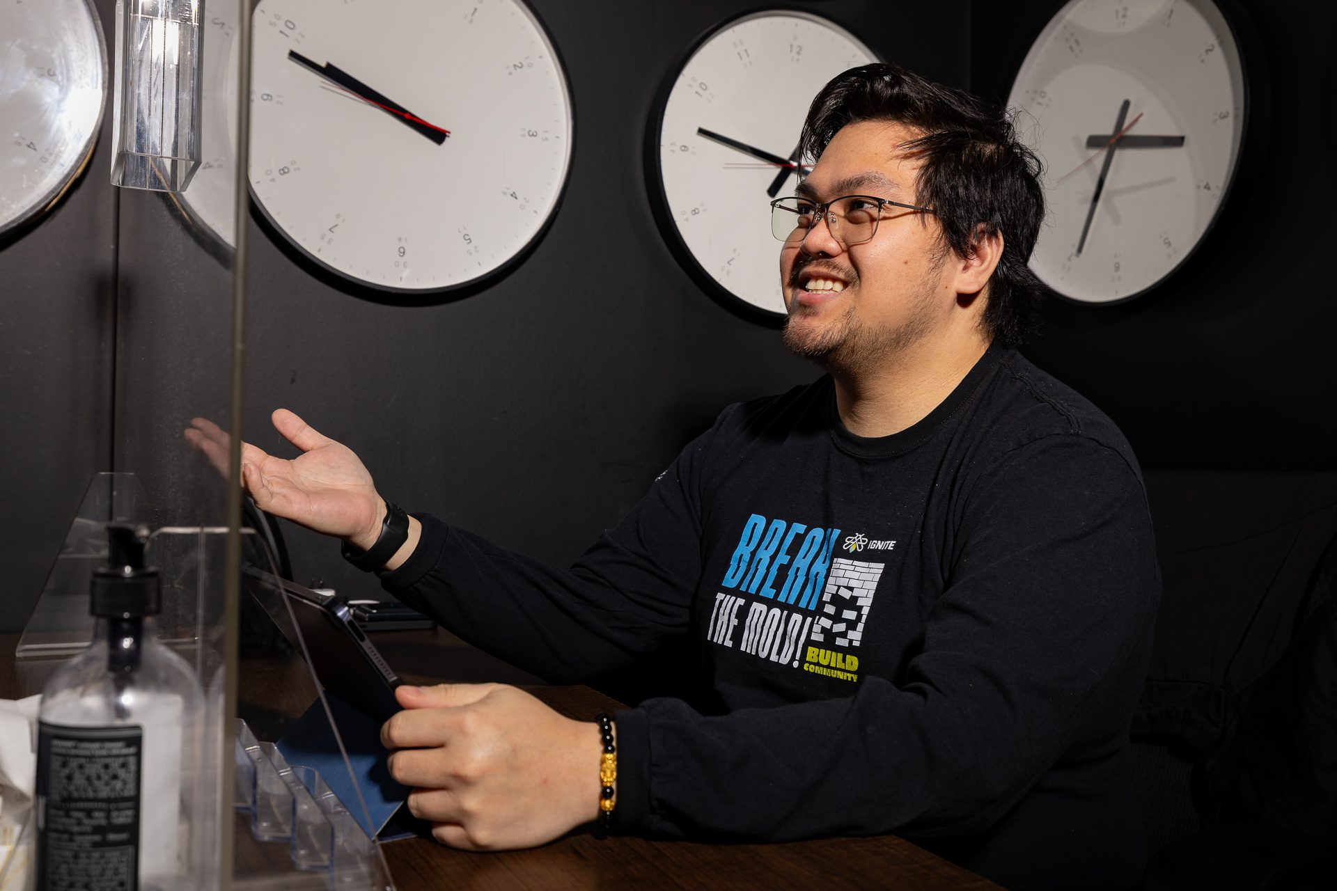 A man with glasses sits at a table, smiling and gesturing with one hand, in front of a laptop. Several wall clocks showing different times are visible behind him. A bottle of hand sanitizer is on the table.