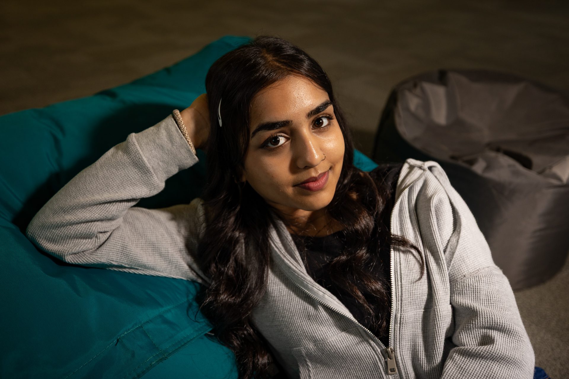 A young woman with long dark hair and a hairpin relaxes on a teal beanbag, wearing a light gray zip-up hoodie. She looks up at the camera with a slight smile, and a gray beanbag is in the background.