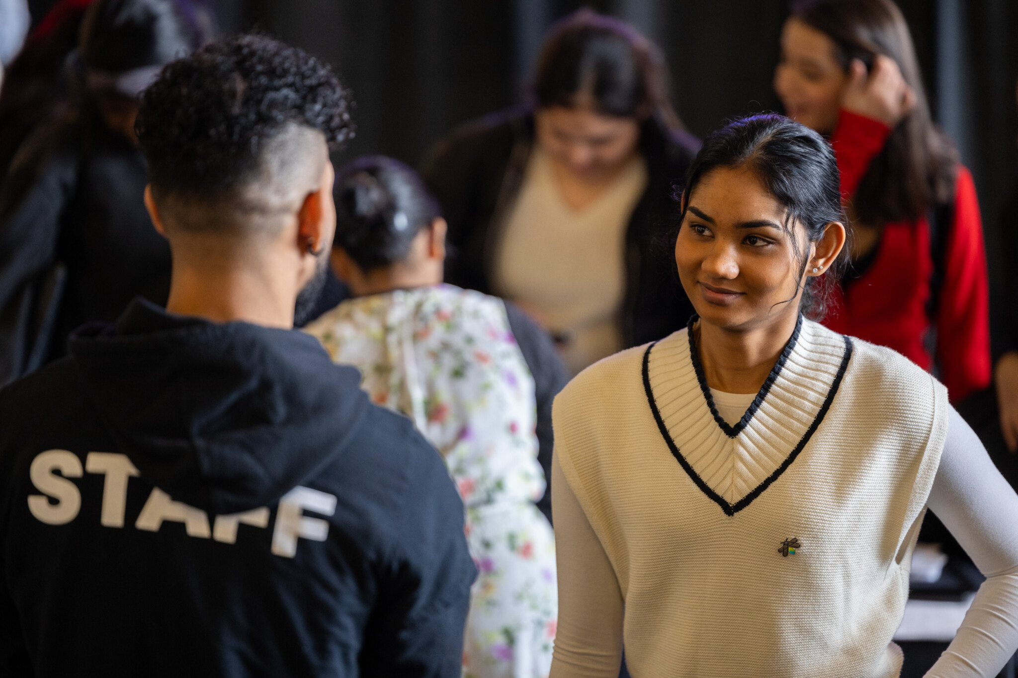 A female student in a sweater vest smiling while conversing face-to-face with an event staff member wearing a black hoodie.