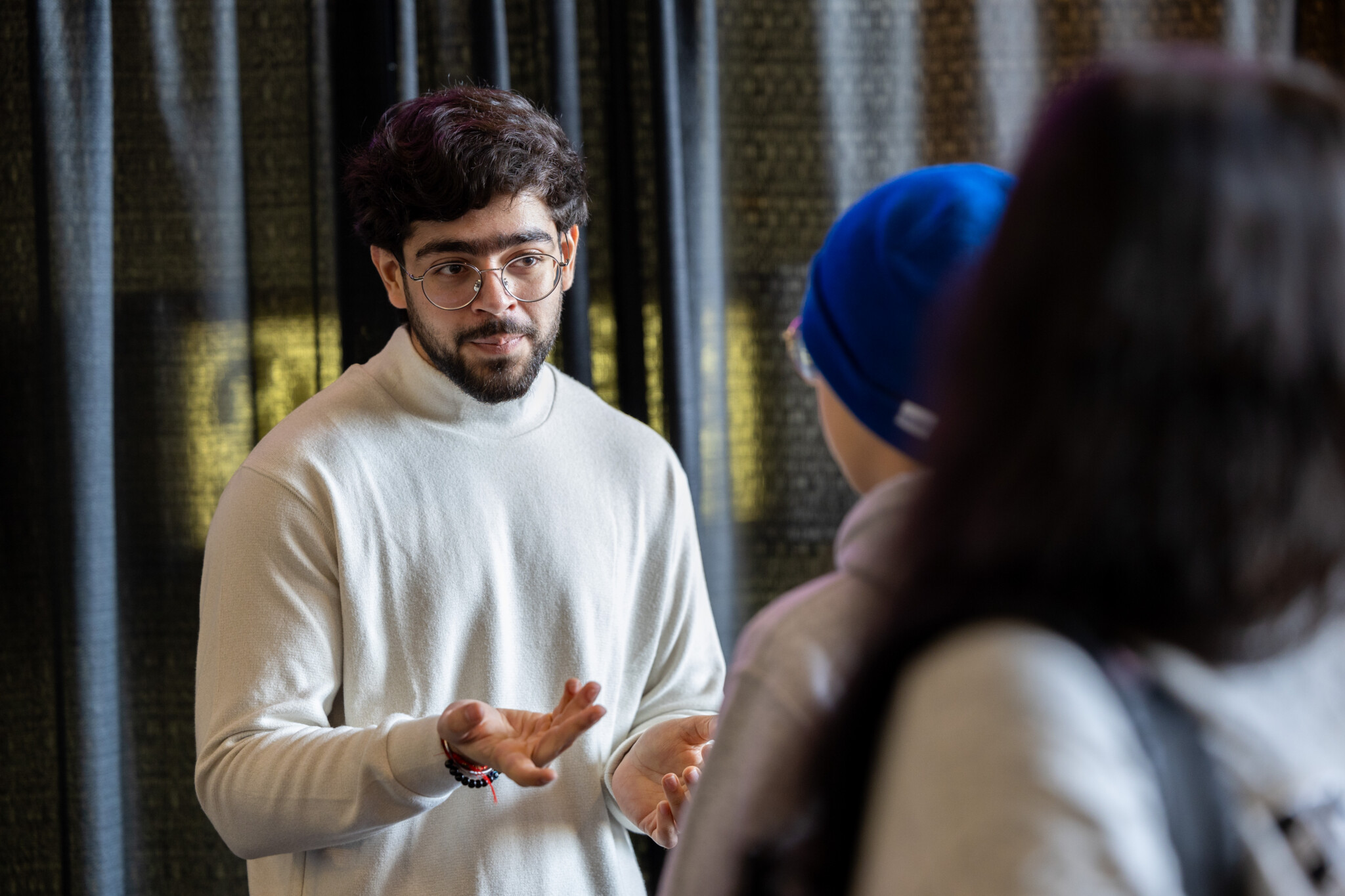 A male student in a cream sweater speaking with open hands to two students at a Meet Your Candidates event.