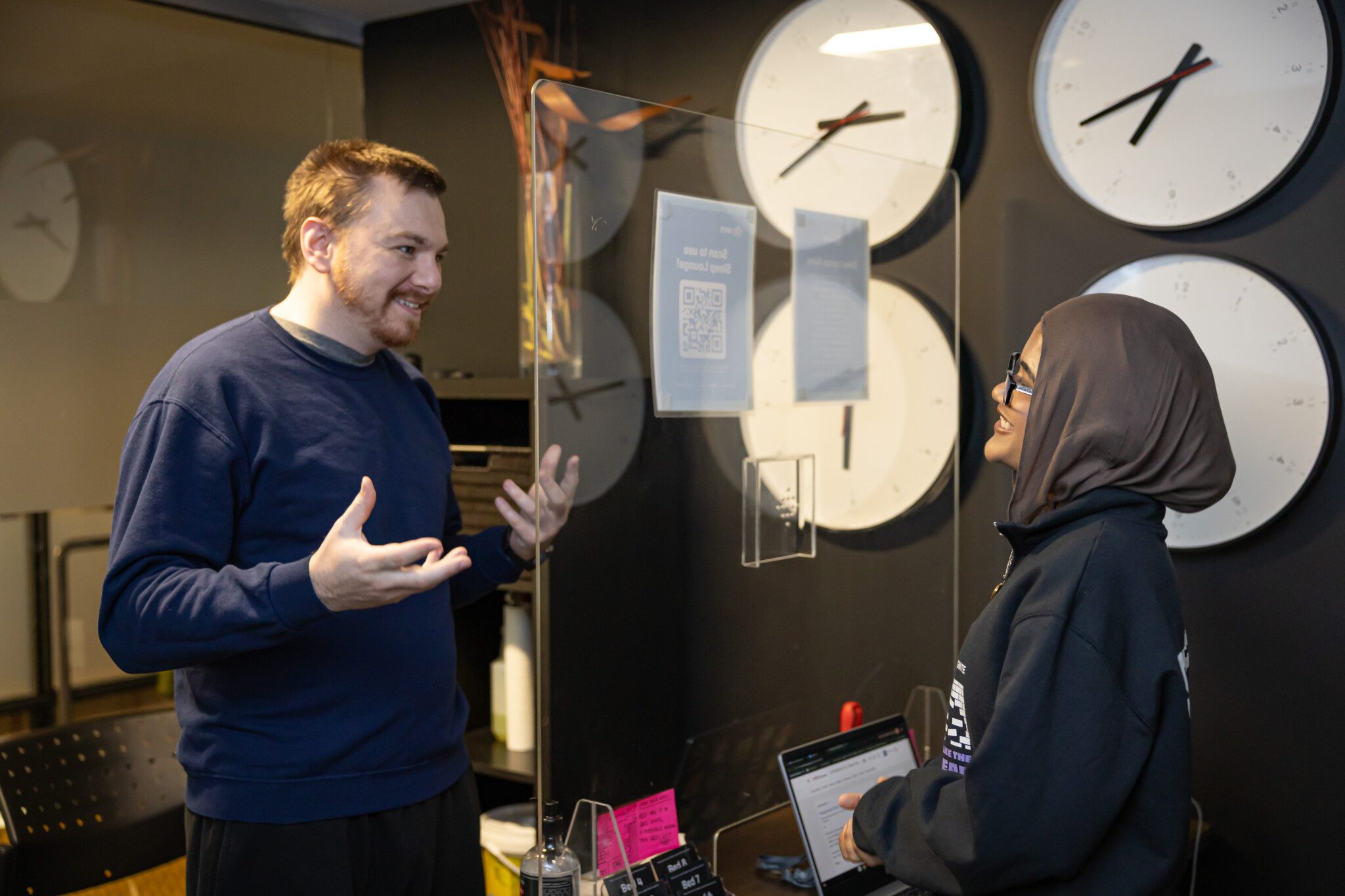 A male student chatting with an IGNITE employee at a reception desk featuring large wall clocks in the background.