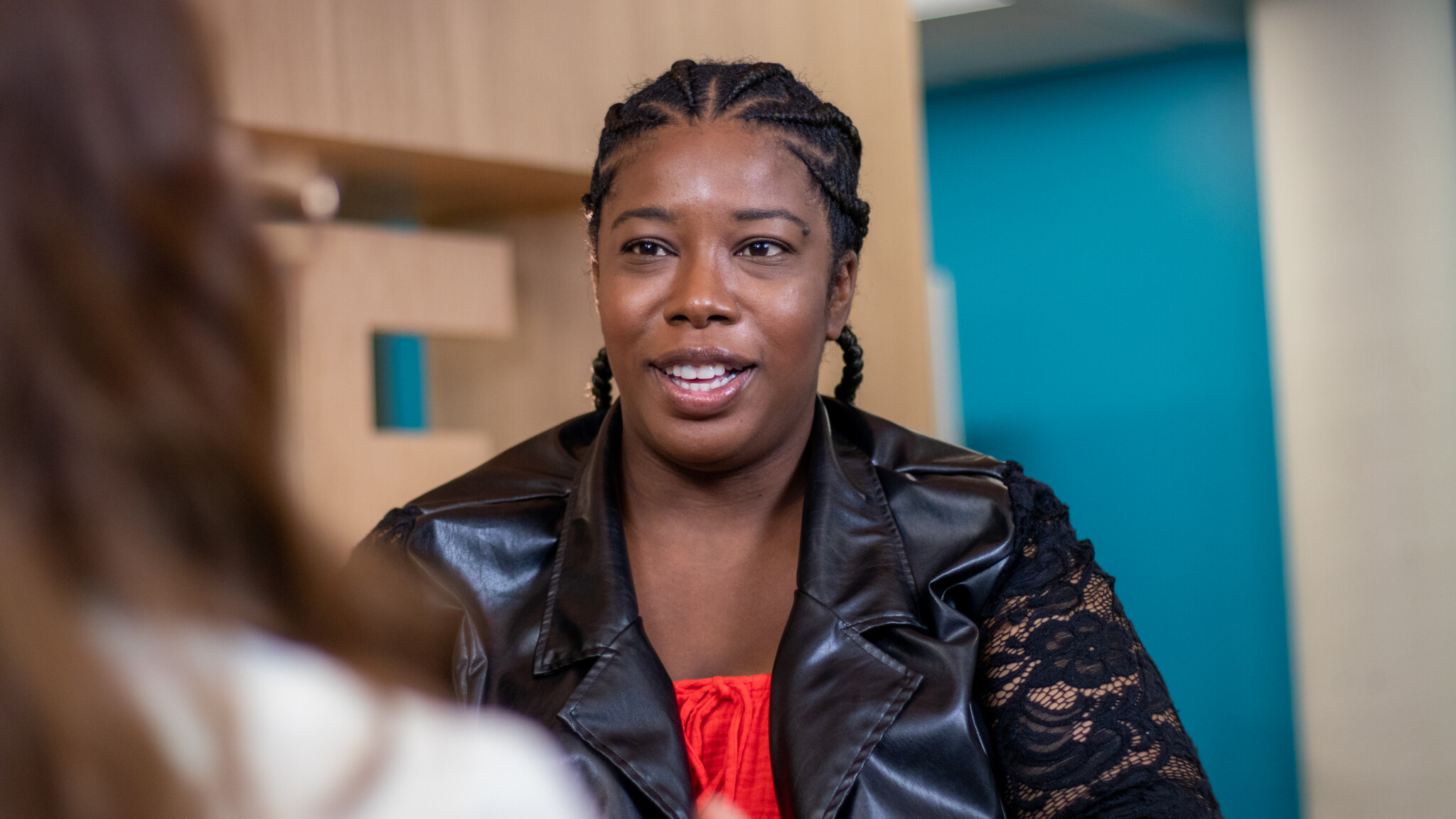 A woman with braided hair, wearing a black leather and lace jacket over a red top, sits indoors and speaks to another person whose back is partially visible in the foreground.