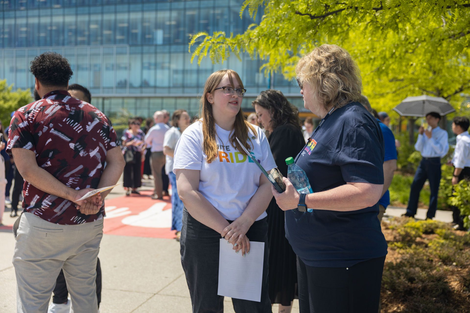 Two women talk outdoors at an event; one wears a PRIDE shirt and holds papers, the other holds a water bottle. People are gathered in the background near a modern glass building.