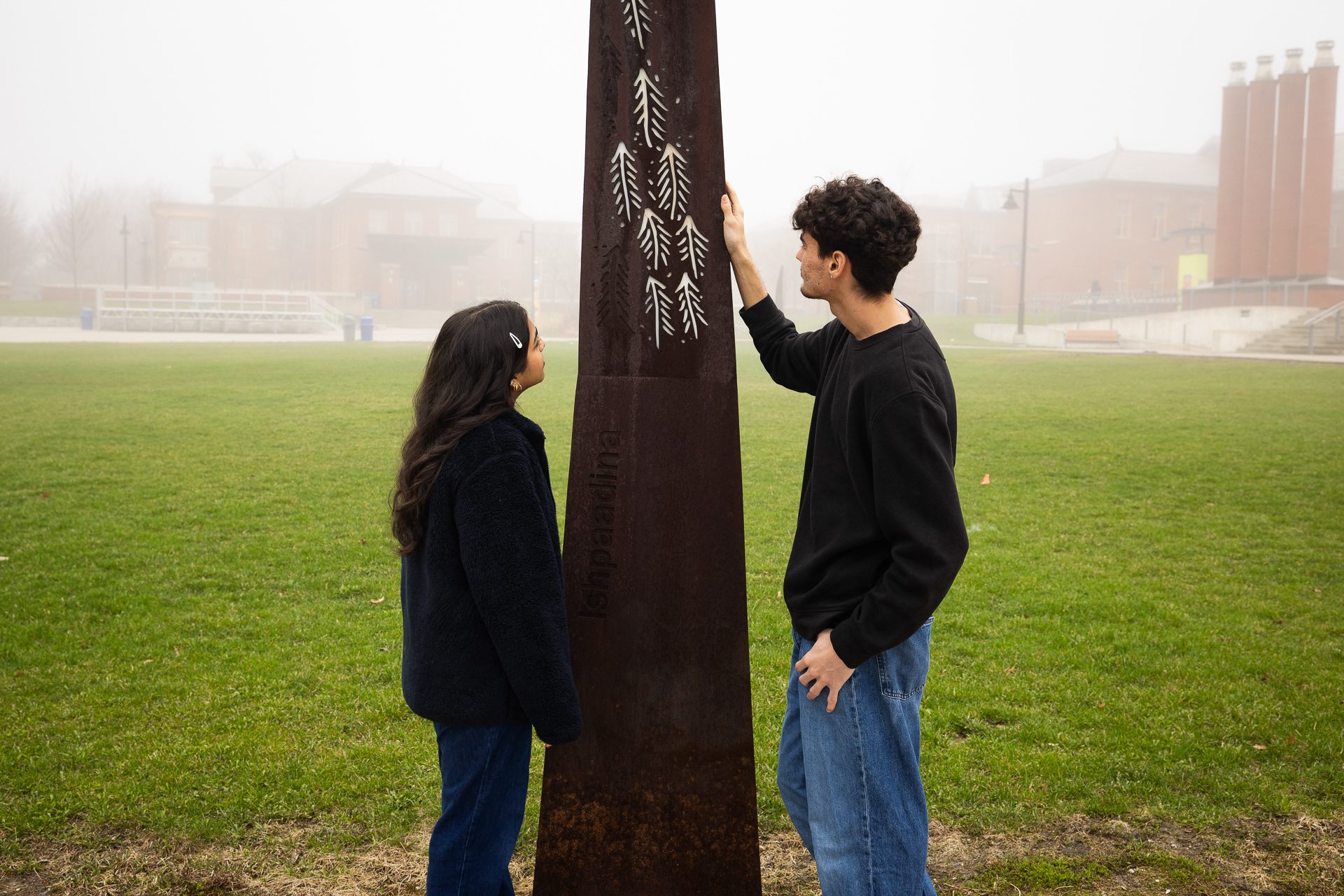 Two people stand outdoors on grass, facing a tall, rust-colored metal sculpture etched with white arrow-like designs. A foggy building and trees are visible in the background.