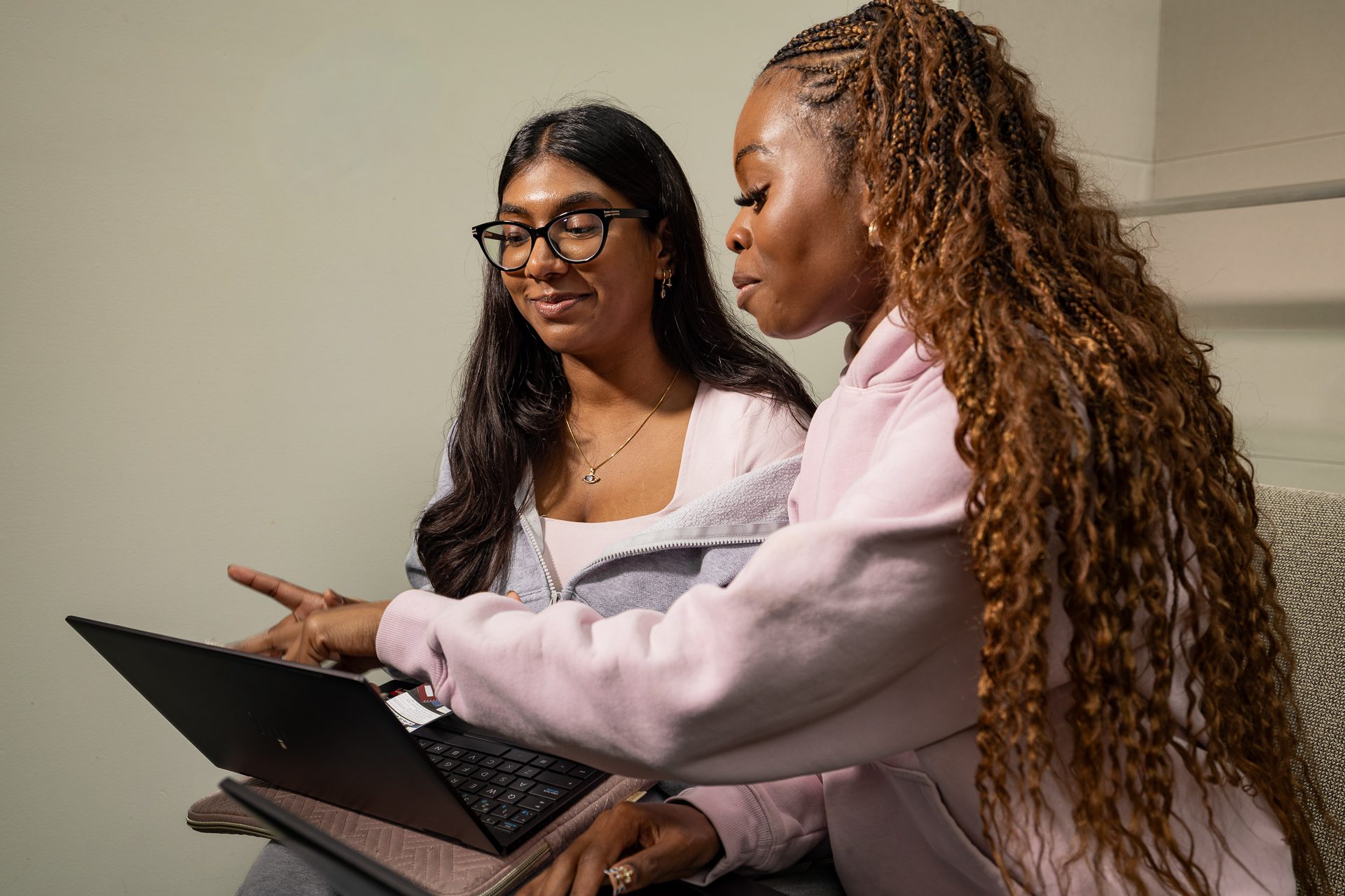 Two women sitting together, one pointing at a laptop screen while the other watches attentively. Both are wearing casual clothes and glasses, and appear to be collaborating or studying.