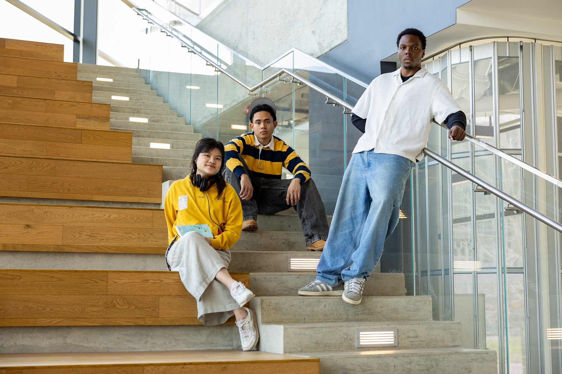 Three young adults pose on a modern wooden staircase indoors; two sit on the steps while one leans against a glass railing, all dressed casually and looking at the camera.