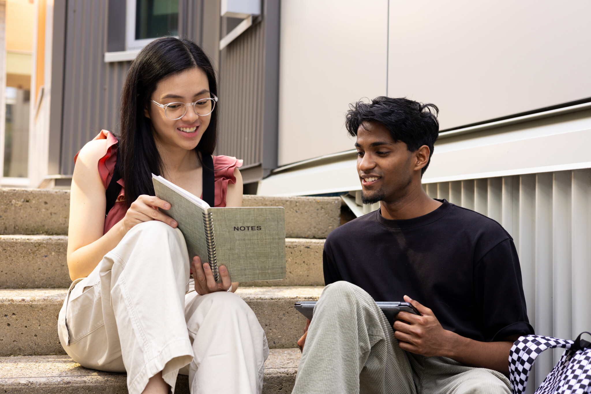 A female student with clear-framed glasses holding a spiral-bound notebook labeled "NOTES" sitting on outdoor concrete stairs alongside a male student in a black t-shirt holding a tablet, both smiling and looking at the notes together.